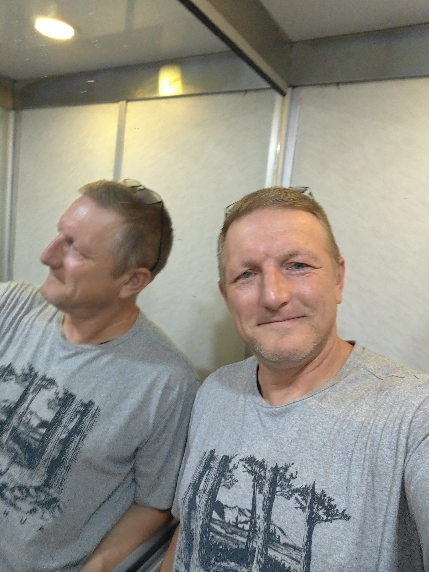 A selfie is taken inside an elevator in the city of Petah Tikva, Israel. The subject, a middle-aged Caucasian male with short blonde hair and a goatee, is wearing a grey t-shirt with a black tree and mountain graphic. He is looking directly at the camera with a slight smile. His reflection is visible in the mirrored wall of the elevator, where he is seen from the side, looking away from the camera. The elevator walls are light-colored and the ceiling has a fluorescent light fixture. There are sunglasses perched on top of the subject's head. The overall environment suggests a utilitarian and enclosed space.