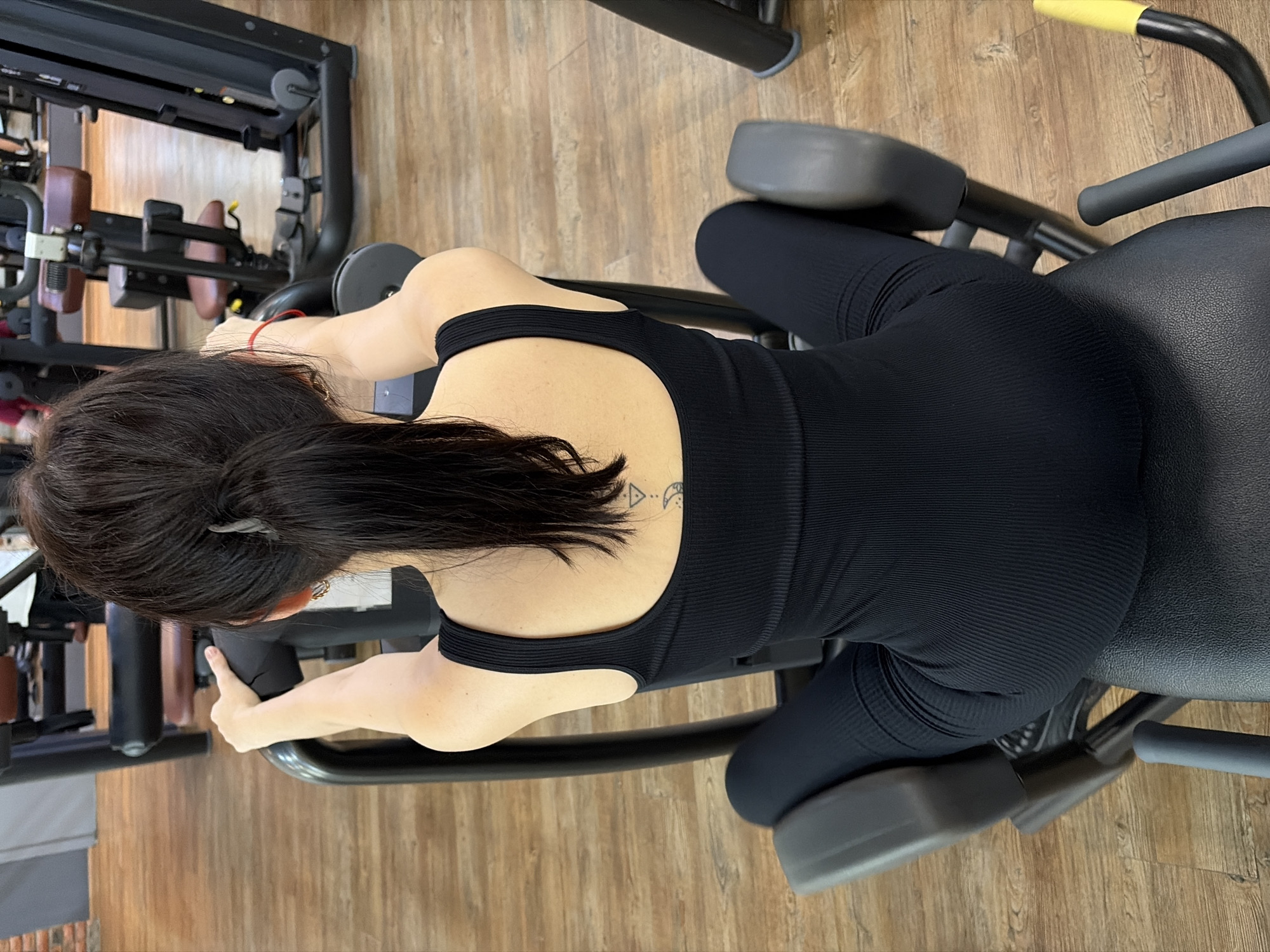 A woman with her back to the camera is seated on a black exercise machine in a gym. She has dark hair pulled back in a ponytail and is wearing a black ribbed tank top and black leggings. A red bracelet is visible on her right wrist. She is holding onto the machine's handles. The gym floor is made of wood. The scene is indoors.