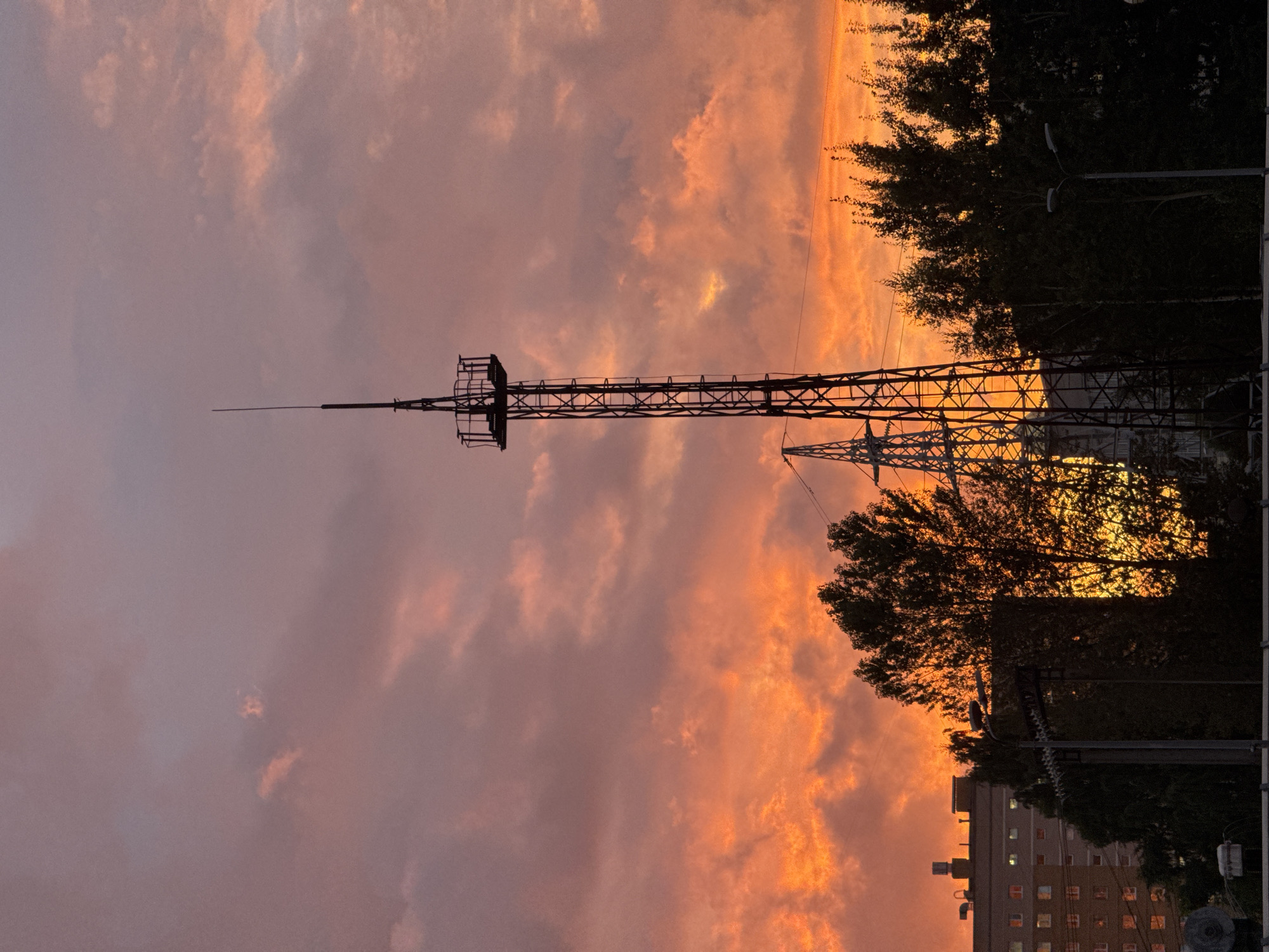 A dramatic sunset paints the sky with vibrant hues of orange and pink, casting a dramatic silhouette over an urban landscape. In the foreground, a tall, lattice-like radio tower stands prominently against the colorful sky, its structure detailed and intricate. To its left, another, shorter tower is partially visible. A dense line of trees forms a dark band across the middle ground, their foliage almost entirely obscured by shadow, with the setting sun creating a bright, almost fiery glow behind them. To the far left, a multi-story apartment building with numerous windows is visible, its lights beginning to twinkle as dusk settles. The overall scene captures a striking contrast between the natural beauty of the sunset and the man-made structures of the city.
