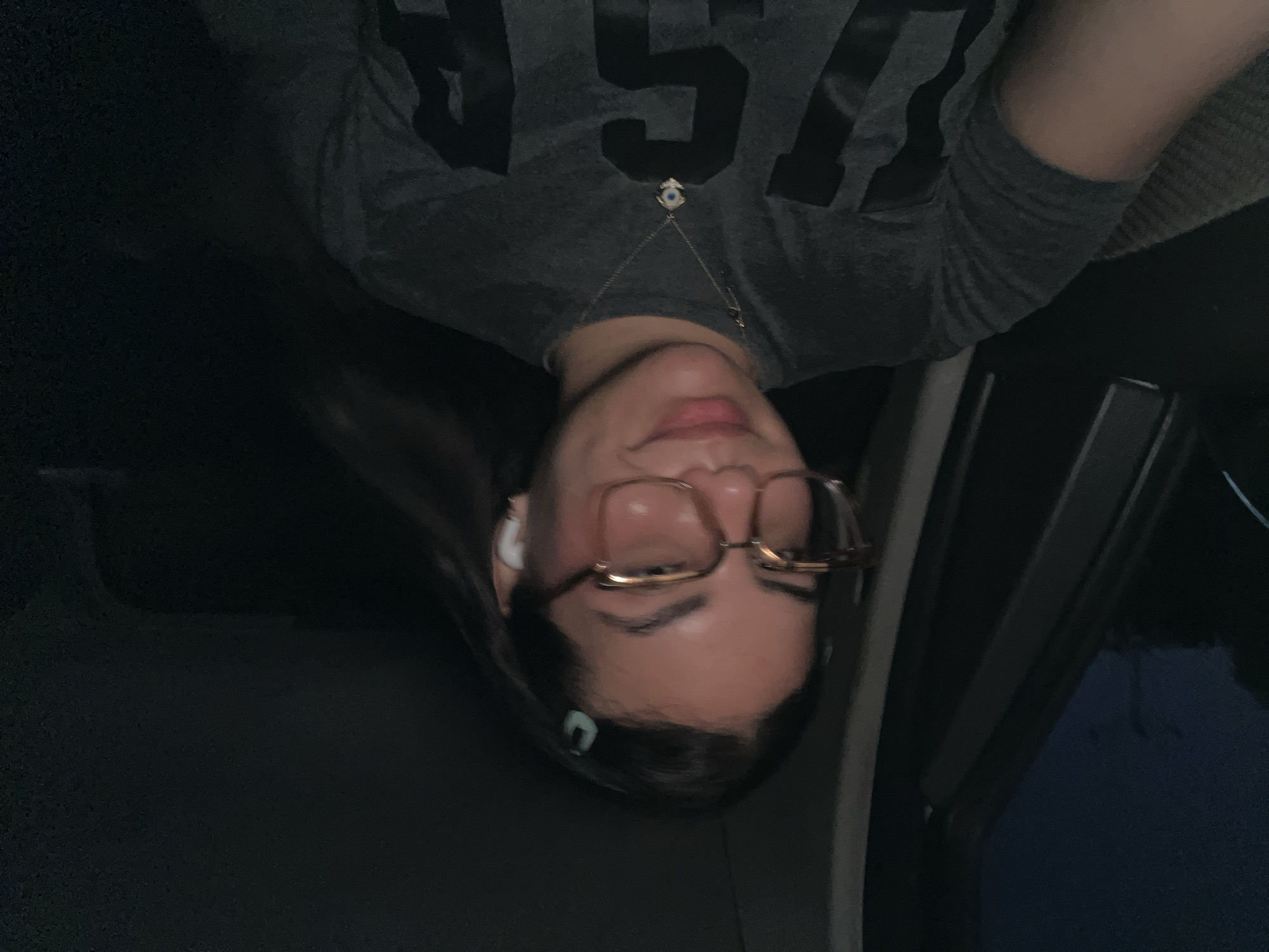 The image shows a close-up, selfie-style portrait of a young woman sitting inside a vehicle at night. She is wearing glasses with thick, square frames and a grey t-shirt with a dark graphic design. A light blue hair clip is visible in her dark hair. She is also wearing white wireless earbuds and a necklace with a blue and white evil eye pendant. The background is dark and out of focus, suggesting it is nighttime. The interior of the vehicle, including part of the dashboard and window, is visible. The scene is set in El Dátil, Venezuela.