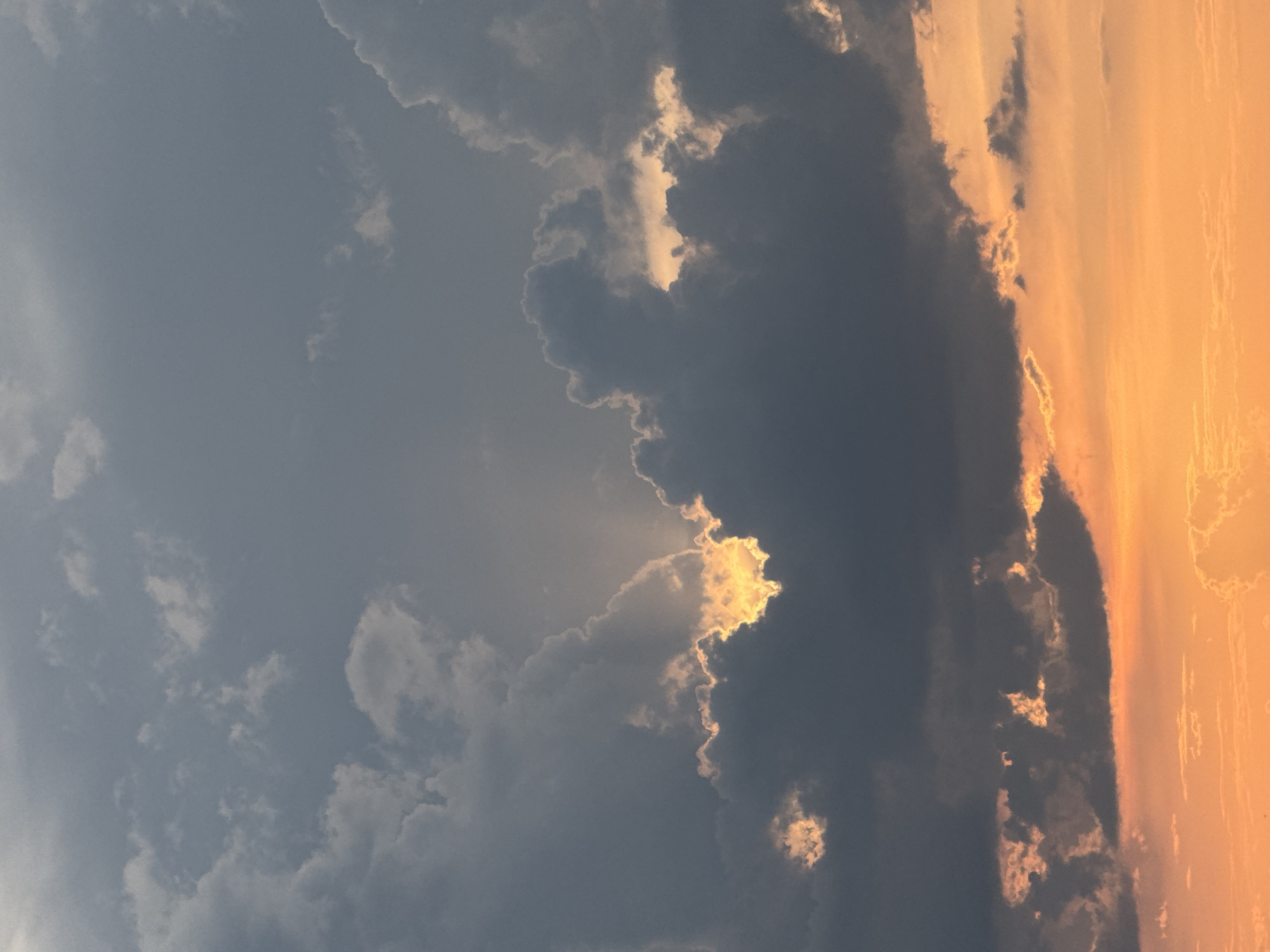 The media file displays an atmospheric sky scene, likely at sunrise or sunset, over Sozopol, Bulgaria. The image is dominated by a dramatic interplay of dark and brightly lit clouds.
On the left side of the frame, the sky is a muted, light grey-blue, with scattered, wispy white clouds. Moving towards the center and right, significant cloud formations become visible. A large, dark, dense cloud mass stretches across the middle of the frame, contrasting sharply with the surrounding light. Within this dark cloud, pockets of intense golden light break through, illuminating the edges and interiors of the clouds with brilliant orange and yellow hues, suggesting the sun is either rising or setting behind or within these clouds. This creates striking outlines and vibrant highlights, particularly in the central and upper-central parts of the image, where rays of light seem to pierce through.
To the right, the sky transitions into a vast expanse of warm orange and golden light, with lighter, streaky cloud formations that are entirely bathed in the rich, fiery glow of the sun. The boundary between the dark clouds and the intensely orange sky is distinct, creating a powerful visual separation. The overall impression is one of dynamic weather and a captivating display of natural light.
There are no people, man-made objects, or discernible activities taking place. No text is visible in the image. The dramatic lighting and warm color palette strongly indicate either dawn or dusk.