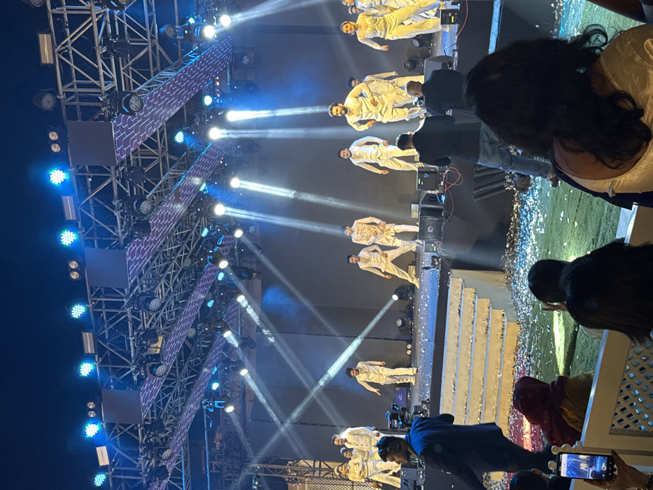 Onstage, performers in white outfits dance under bright spotlights. A scaffold with blue-lit squares and patterns supports lighting equipment. Confetti litters the stage floor. Audience members, including some holding phones, watch the show. The view is slightly obstructed.