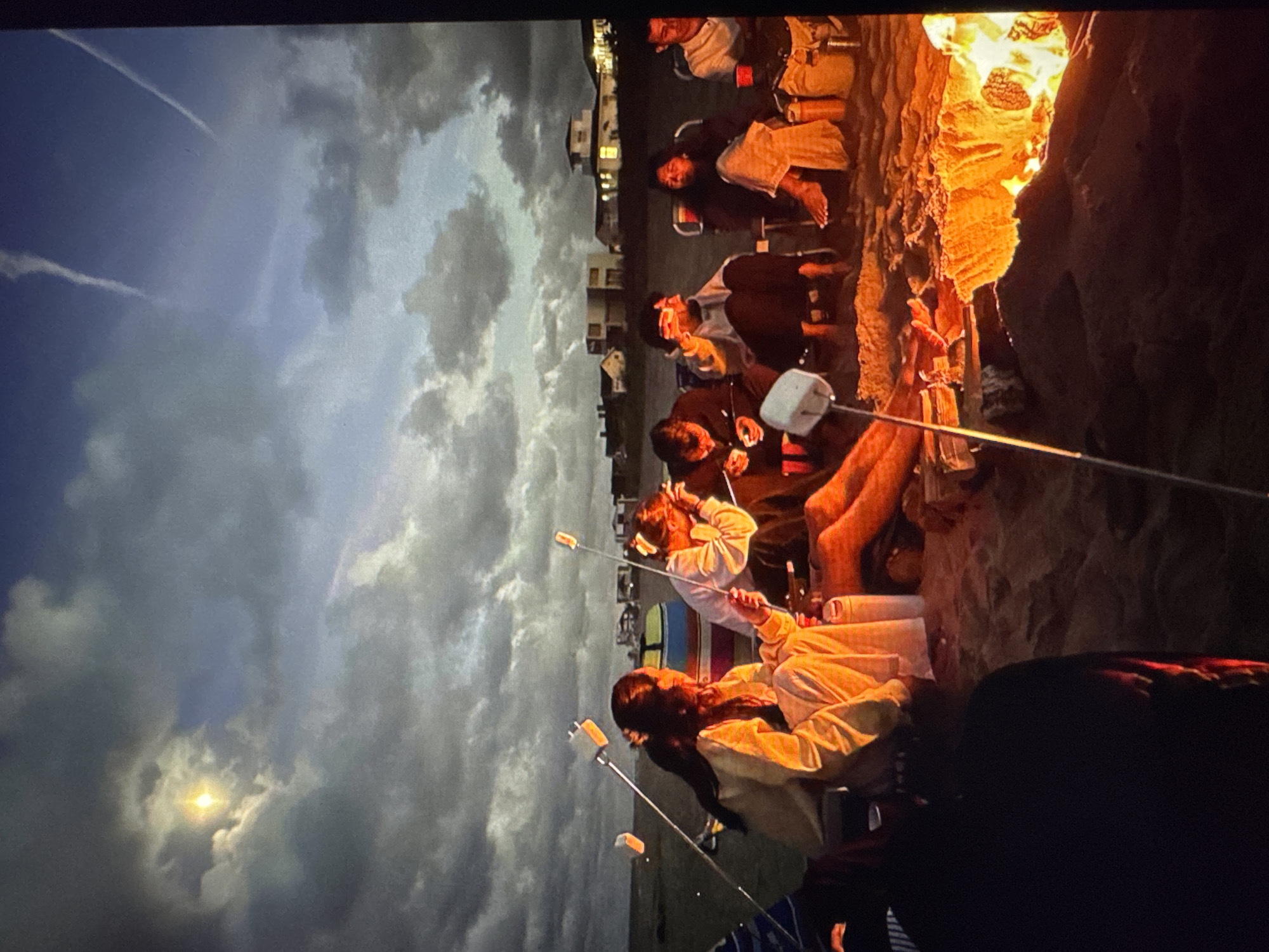 The image depicts a nighttime beach scene with a group of people gathered around a bonfire. The individuals are roasting marshmallows on sticks over the flames. The sky is dark and cloudy, with a bright moon visible through a break in the clouds. In the background, there are houses and what appear to be recreational vehicles or tents. The setting is a sandy beach. The overall atmosphere is casual and communal. The image is a photograph of a group of people on a beach at night, around a bonfire. The people are roasting marshmallows. In the background, there are houses and what looks like a campground or RV park. The sky is cloudy, but the moon is visible.