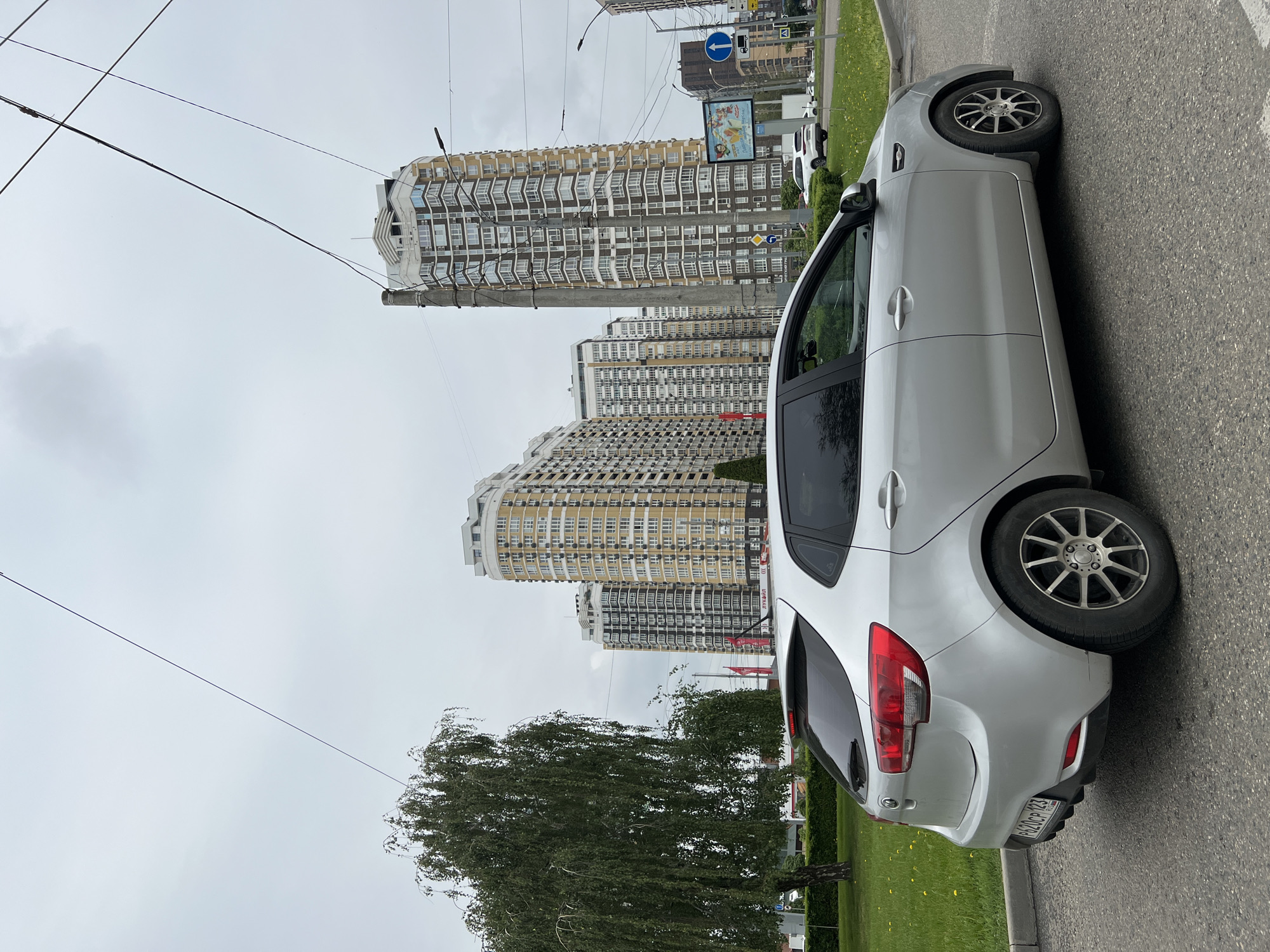 A silver Kia Rio is parked on the side of a road in what appears to be Krasnodar, Russia, based on the license plate number. The rear of the car is visible, including the taillights, bumper, and license plate. Tall, multi-story apartment buildings with cream and brown facades are in the background. A green grassy area with a tree is between the car and the buildings. Overhead power lines run across the sky. The sky is overcast. A road sign is visible in the background near the top of the frame.