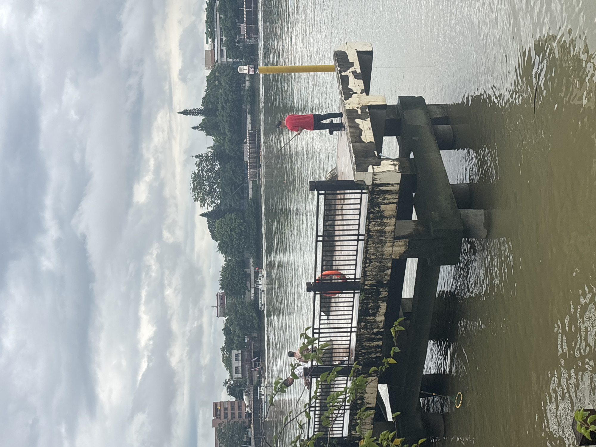 In the Nanhai District of China, an overcast day unfolds by a wide, murky river. A concrete pier extends into the water, its surface weathered and stained. On this pier, two individuals are engaged in fishing. One person, wearing a red shirt and dark pants, stands with a fishing rod, casting into the water. Another person, dressed in lighter clothing, is positioned further back on the pier, also holding a fishing rod. A black metal railing lines the edge of the pier, with an orange life preserver attached to one of its posts. Supporting the pier are large, moss-covered concrete pillars submerged in the river. The riverbanks are lined with lush green trees, and buildings are visible in the distance, interspersed with the foliage. The sky is heavily clouded, creating a subdued lighting condition over the scene.