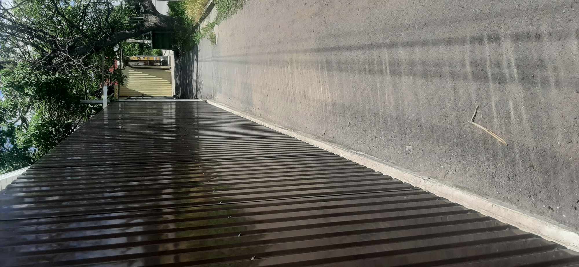The image shows a narrow asphalt path running alongside a tall, dark brown corrugated metal fence. The fence occupies the left two-thirds of the frame and is in sharp focus. The path extends to the right, receding into the background. A small twig lies on the asphalt path in the foreground. In the background, to the right of the fence, is a building with a yellow corrugated metal exterior. Beyond the building, there are trees with green foliage and a clear sky. The scene appears to be an outdoor alleyway or path within a residential or commercial area.