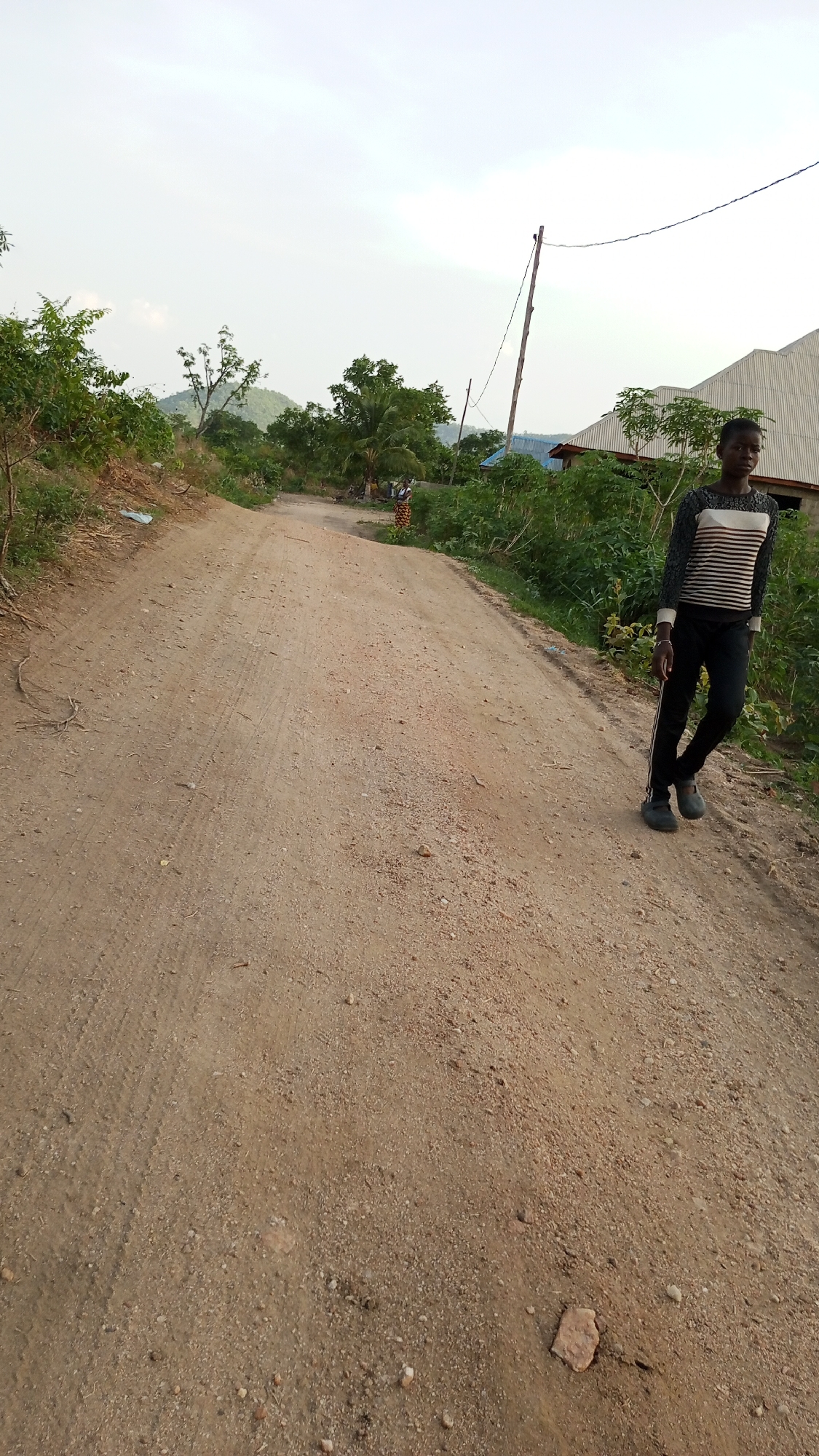 A dirt road dominates the foreground of the scene, its surface marked by tire tracks and scattered debris. The road leads into the background where two people are walking. To the right of the road, a young person stands wearing a horizontally striped long-sleeved shirt, black pants, and dark shoes. Houses and trees line the road, and an electrical pole is visible near a house. The sky is overcast. The location is the city of Adankolo, Nigeria.