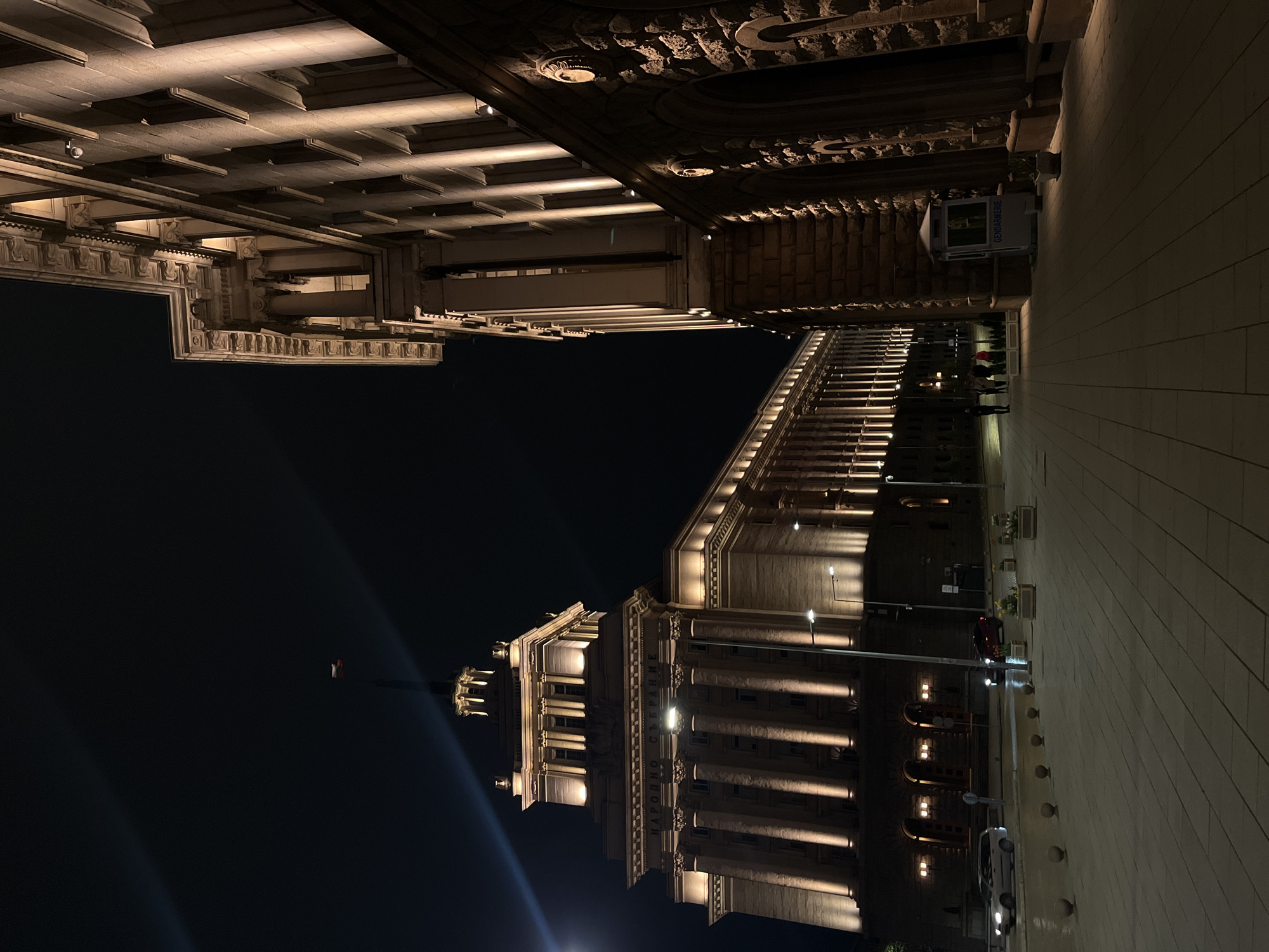 This nighttime image captures a grand, classical-style building, confirmed by the visible text "НАРОДНО СЪБРАНИЕ" (National Assembly) on its facade, indicating the National Assembly of Bulgaria in Sofia. The architecture features numerous tall columns, ornate cornices, detailed capitals, and sculpted reliefs, all illuminated by warm-toned floodlights and sconces. The building appears to be constructed from light-colored stone or concrete.
To the right, another similarly styled building, possibly part of the same complex or a neighboring structure, stretches into the background. The sky is dark, emphasizing the artificial illumination. On the ground level, a wide, paved plaza or street is visible, with several streetlights casting beams across the scene.
A few small figures, appearing to be people, are seen walking or standing on the ground near the buildings. A small white booth with "GENDARMERIE" printed on its side is visible, suggesting a security presence. Further along the street, a dark-colored car is parked, and a white car is visible in the distance. Planters with flowers add small touches of greenery. The overall atmosphere is quiet and stately, highlighting the architectural grandeur under the night sky.