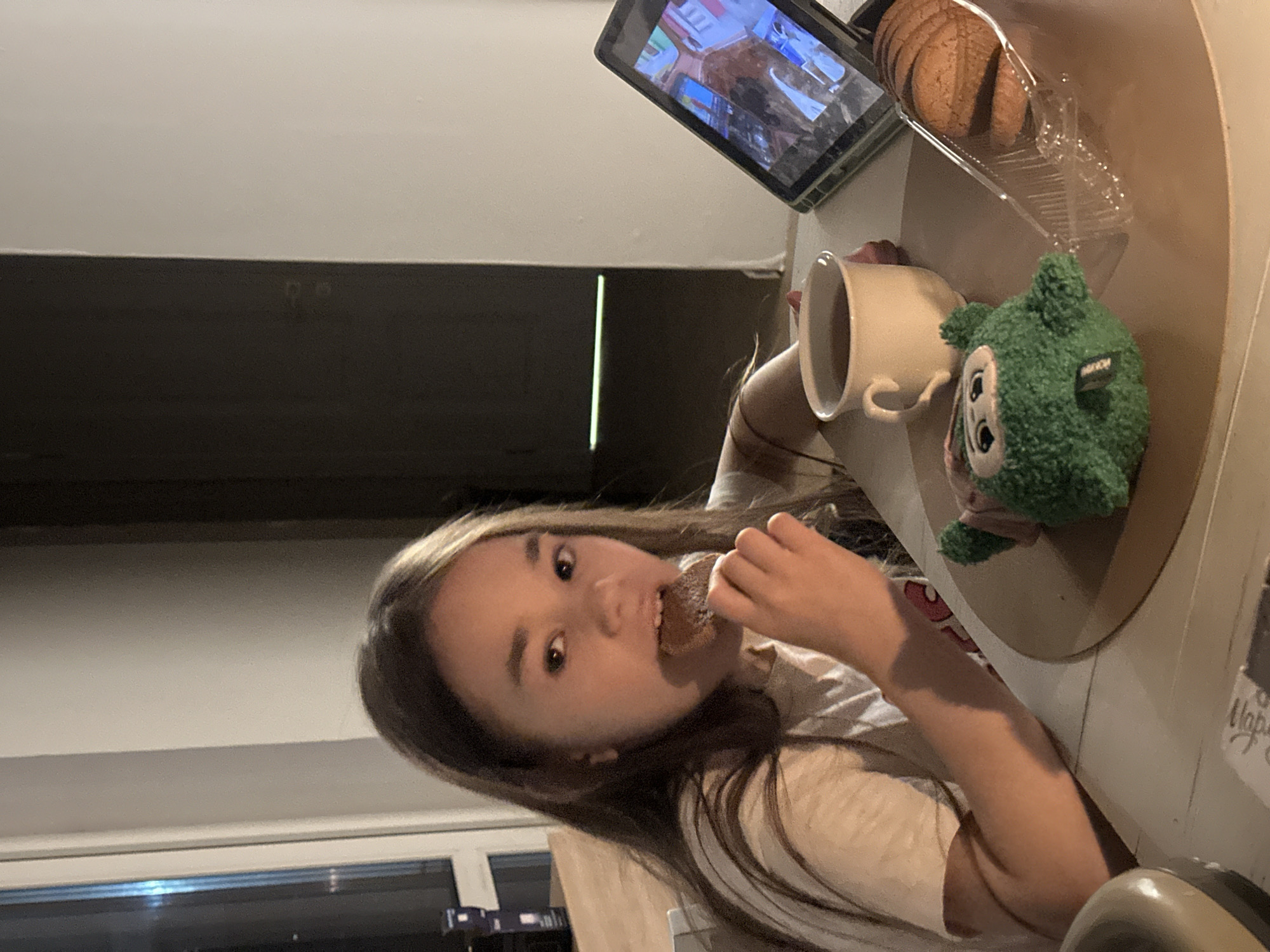 A young girl is seated at a table in an interior setting, possibly a home. She is holding and about to eat a dark-colored cookie. A tablet is positioned on the table, displaying an unidentified video. To the right of the tablet are a container of cookies, a cup, and a stuffed green monster toy. The background includes a window and what appears to be a cabinet. There is no evidence to place the location in Kharnavy, Russia.