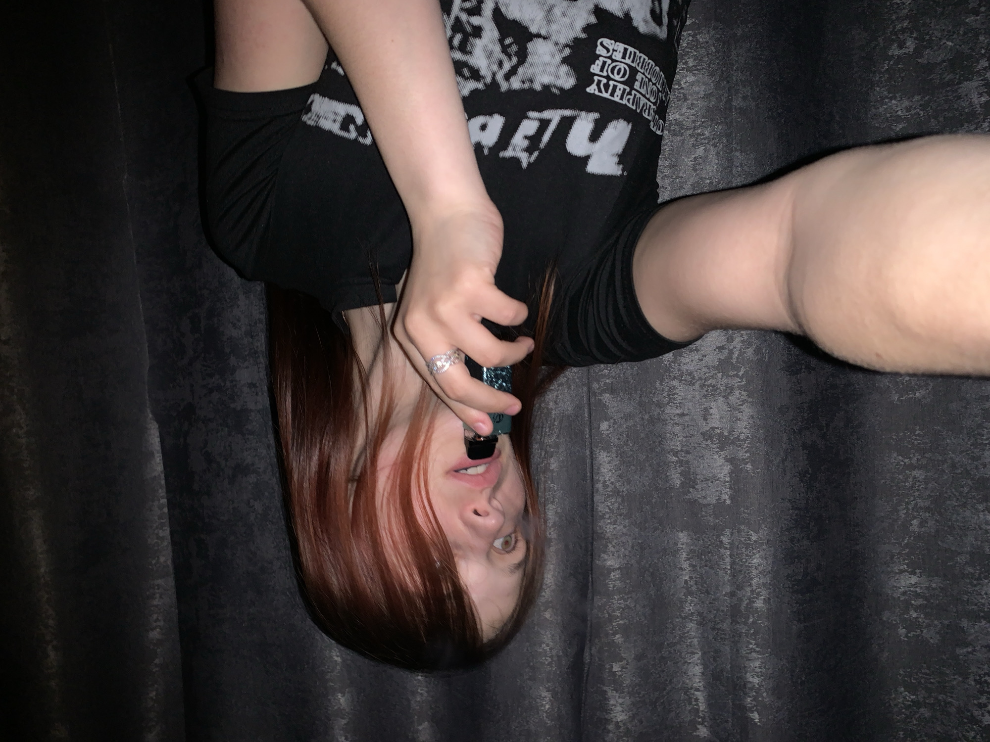 A person is positioned upside down, holding a dark-colored object to their mouth with their right hand. They are wearing a black shirt with a white design on the front and black shorts. The person has red hair, fair skin, and is wearing a ring on the ring finger of their right hand. The background consists of dark gray curtains. The image does not provide enough details to identify the location in the city of Фоки, Russia.