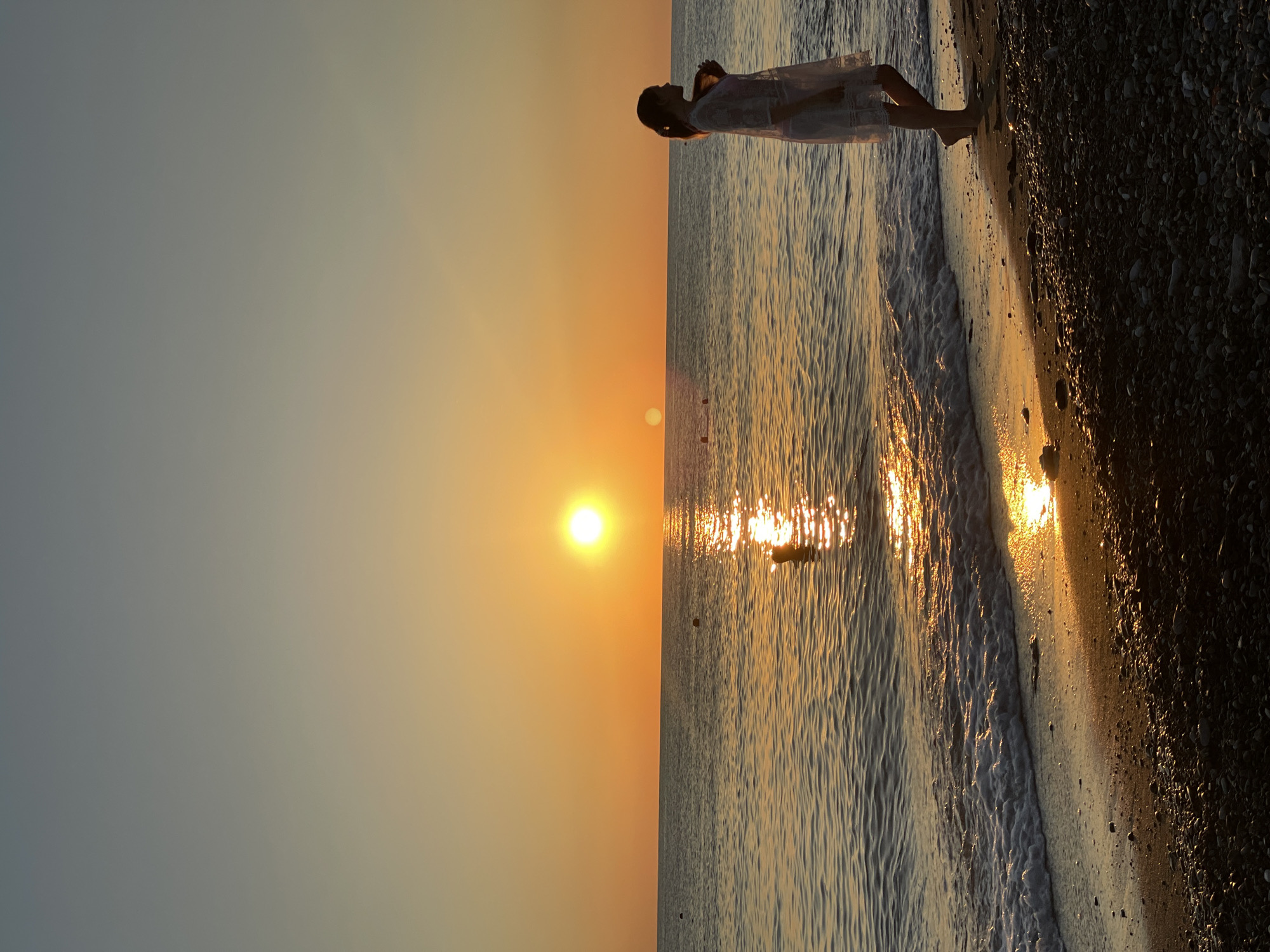 A woman stands on a pebble beach at the edge of the ocean, silhouetted against a setting sun. The sun is a bright orange disc low on the horizon, casting a golden reflection across the water. Gentle waves lap at the shore, creating a foamy white line where the water meets the sand and pebbles. The sky is a gradient of orange and yellow near the horizon, fading into a soft blue-grey at the top of the frame. The woman, seen in profile, is wearing a light-colored dress or cover-up over a swimsuit. Her hair is pulled back. In the distance, a few buoys or markers are visible in the water, and a person is partially submerged in the sea. The beach itself is covered in small, dark pebbles and some sand. The overall mood of the image is serene and peaceful, capturing a moment of quiet reflection by the sea.