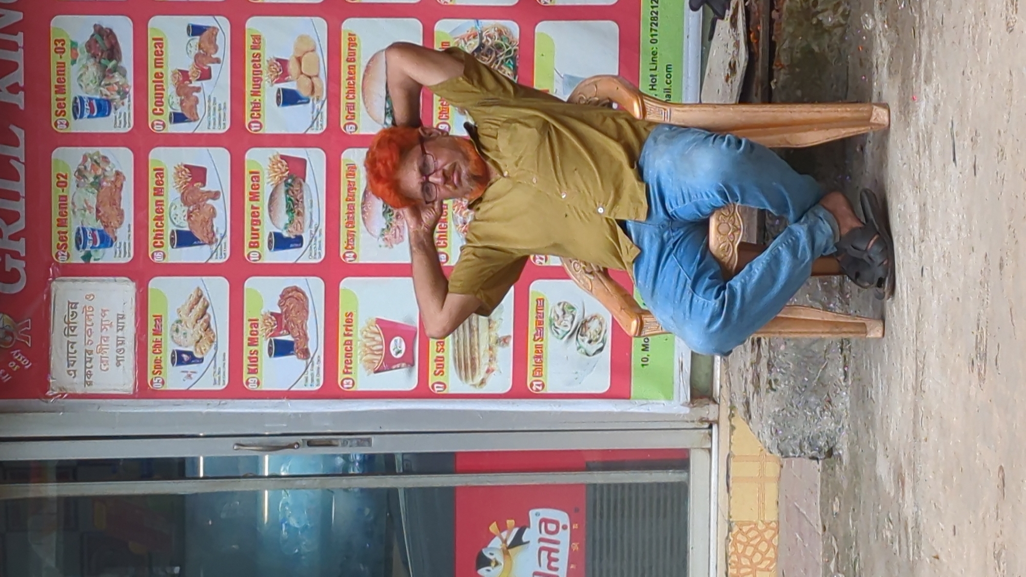 A man with bright orange hair and beard, wearing glasses, a mustard-colored shirt, and blue jeans, sits in a plastic chair outside a fast-food restaurant. His hands are behind his head. The restaurant's menu is displayed on a large board behind him, featuring various food items like chicken meals, burgers, and fries. Some of the menu items are in English, while others are in Bengali. The restaurant appears to be located in Dhaka, Bangladesh, as indicated by the Bengali text on the menu and a visible address "10, Mo..." which likely refers to a road or area in Dhaka. The environment is urban, with a concrete sidewalk and a shop window to the left.