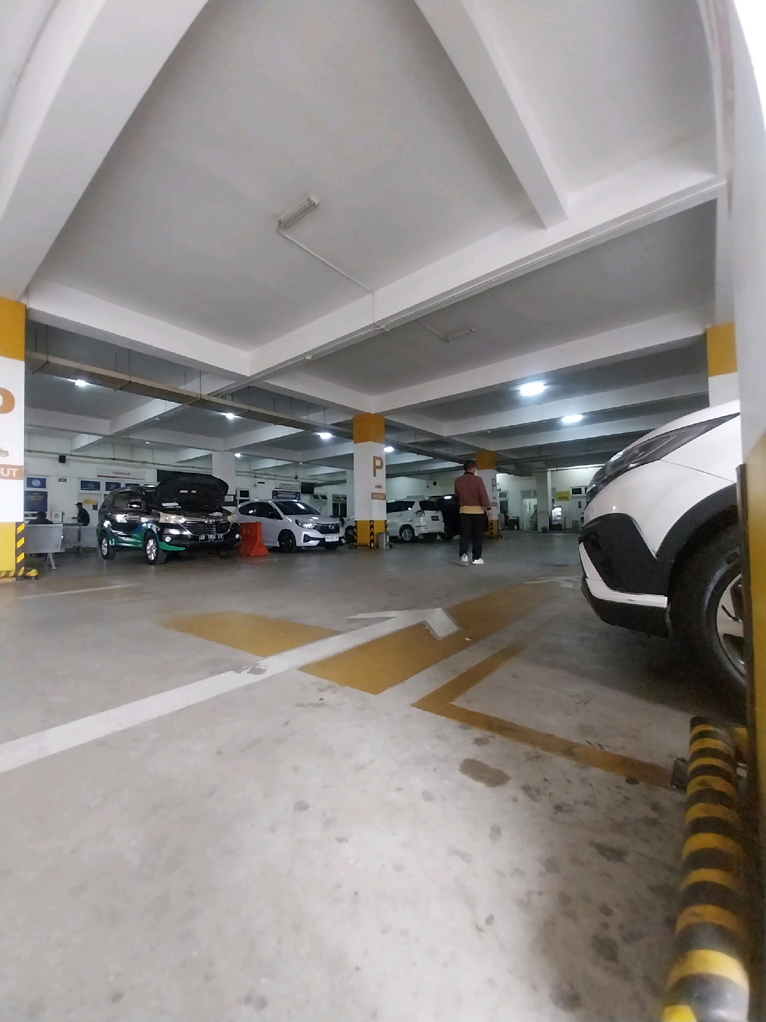 A wide-angle, eye-level shot captures an interior parking garage in Sleman, Indonesia. The concrete floor is marked with yellow and white directional lines. Several cars are parked, including a black SUV with its hood open, a white sedan, and a white hatchback in the background. A person in a brown jacket and black pants walks away from the camera towards the left. The ceiling of the garage features exposed beams and fluorescent lighting. Yellow and black striped bollards are visible along the right edge of the frame. A sign with the letter "P" on a yellow background is mounted on a pillar.