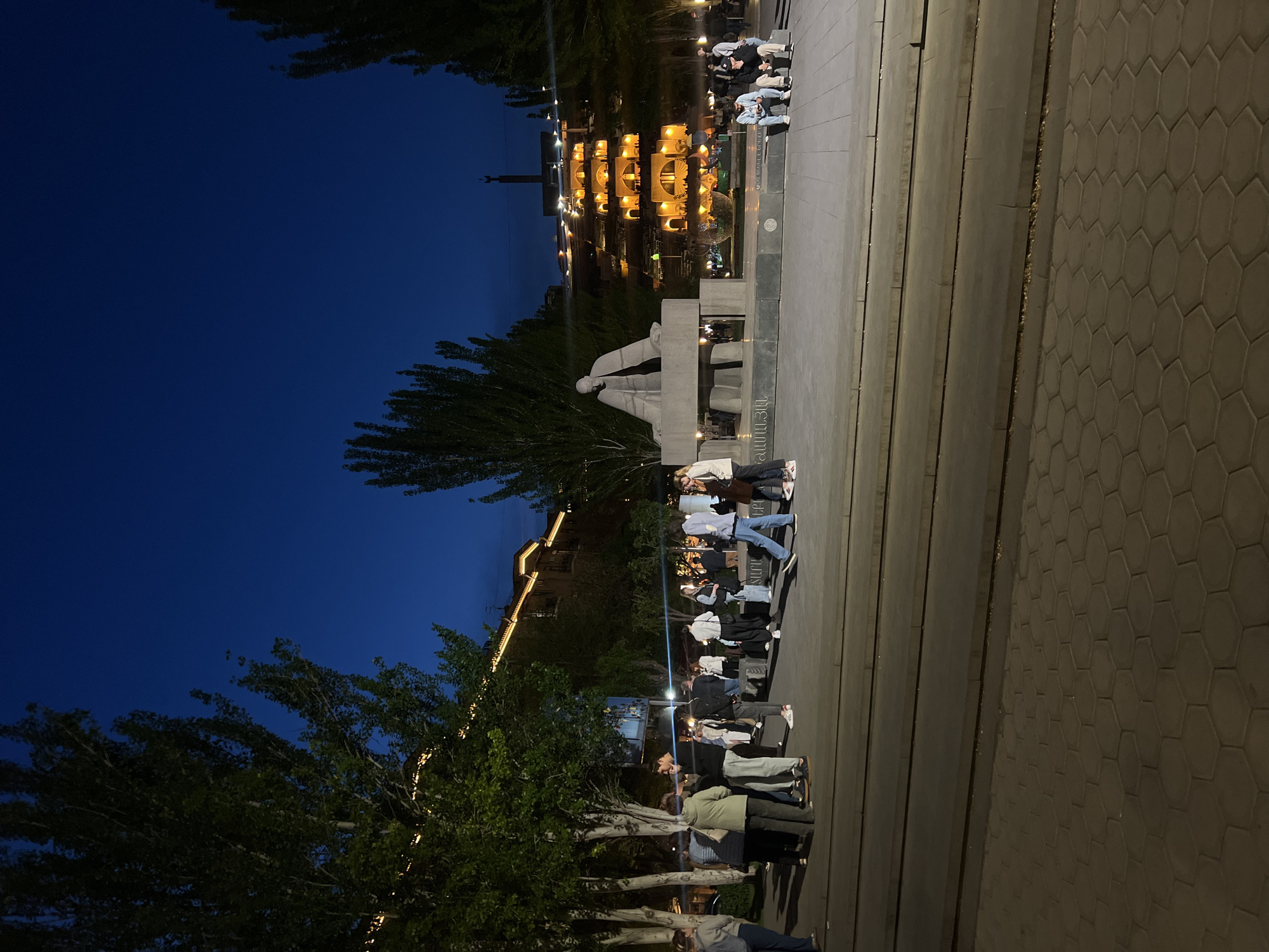 The image shows a nighttime scene in Yerevan, Armenia, likely at the Cascade complex area. A gray monument sits in the foreground beneath the Cascade. People stand along the sides of the Cascade levels. Some individuals are seated. The environment is well-lit, with lights illuminating the Cascade structure and surrounding areas. The sky is a deep blue. Trees are visible on either side of the frame. A building with illuminated archways is situated in the background. The ground is paved with gray stones.