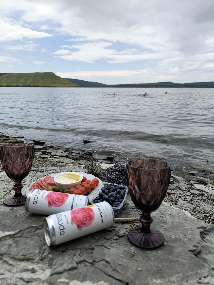 The image depicts a riparian scene near Horaivka, Ukraine, likely along the Dniester River or a reservoir. In the immediate foreground, a rocky shoreline composed of flat, greyish-brown stones serves as a surface for a picnic arrangement. This arrangement includes a white, scallop-edged plate holding circular slices of cured meat, a small white bowl containing a yellow substance (e.g., butter or cheese spread), several red cherry tomatoes, and two green gherkin pickles. Adjacent to the plate is a clear plastic container filled with blueberries. Two purple-tinted, diamond-patterned glass goblets are positioned on the rocks, flanking the food. Two white aluminum cans, labeled "sprag" and "комбуча" (kombucha) with pink fruit illustrations, lie near the plate, one upright and one tilted. A small bundle of dried purple flora, possibly lavender, is visible behind the plate.
The mid-ground features a wide expanse of rippled water. Within the water, three human figures are discernible, partially submerged, suggesting swimming or wading activity. The distant background presents a landscape of rolling green hills, indicative of a river valley. The sky above is partially cloudy, displaying both patches of light blue and areas of grey cloud cover. The overall illumination is consistent with daylight conditions.