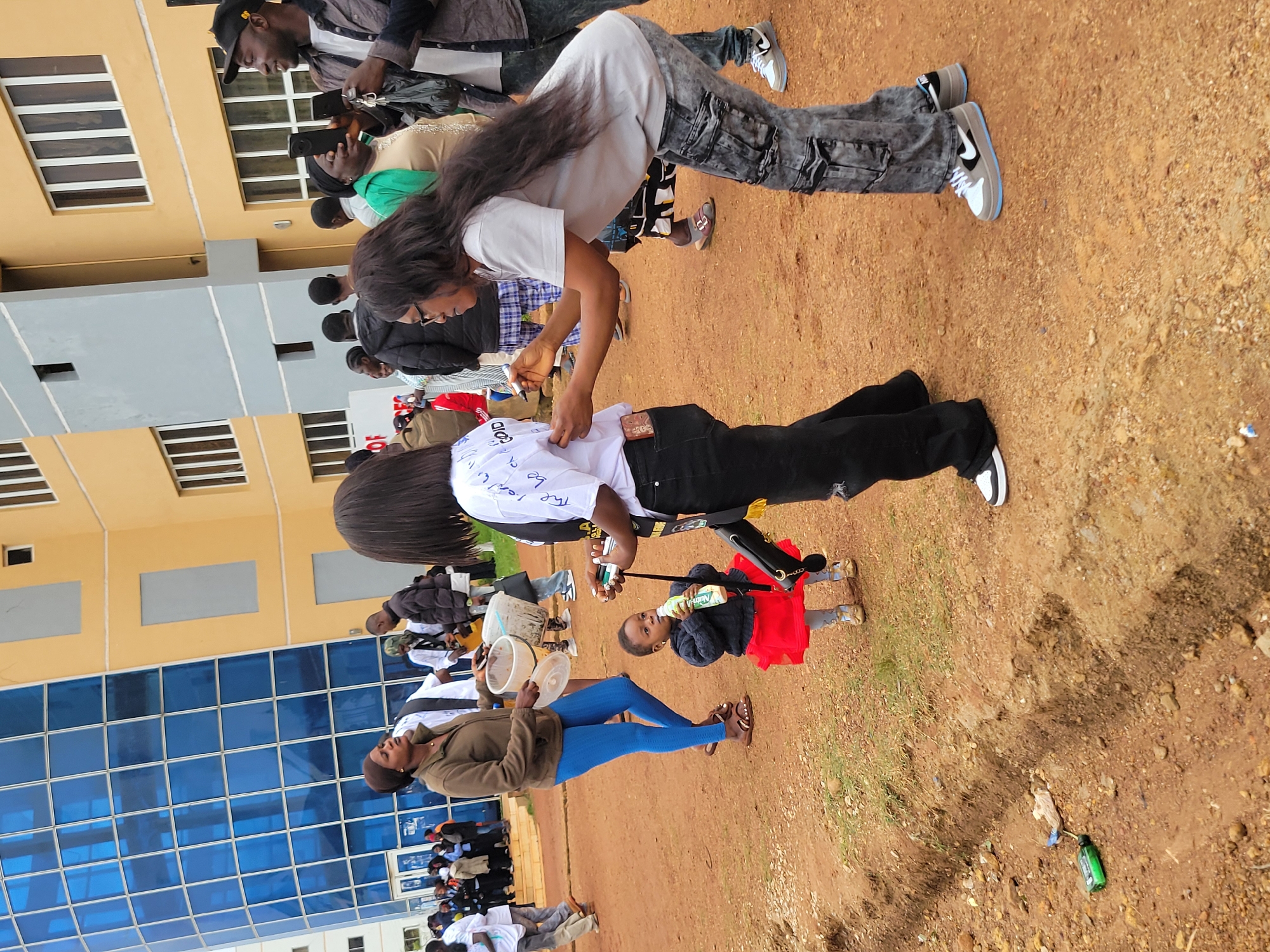 In Durumi, Nigeria, a group of people are gathered outside a modern building with a blue-tinted glass facade and yellow accents. In the foreground, a woman in grey is signing the back of a white t-shirt worn by another individual. Beside them, a young child in a red skirt and dark top is drinking from a bottle. A woman in blue leggings and a brown jacket holds two white plastic containers. Other individuals are visible in the background, some holding phones and interacting with each other. The ground is a mix of dirt and sparse vegetation.