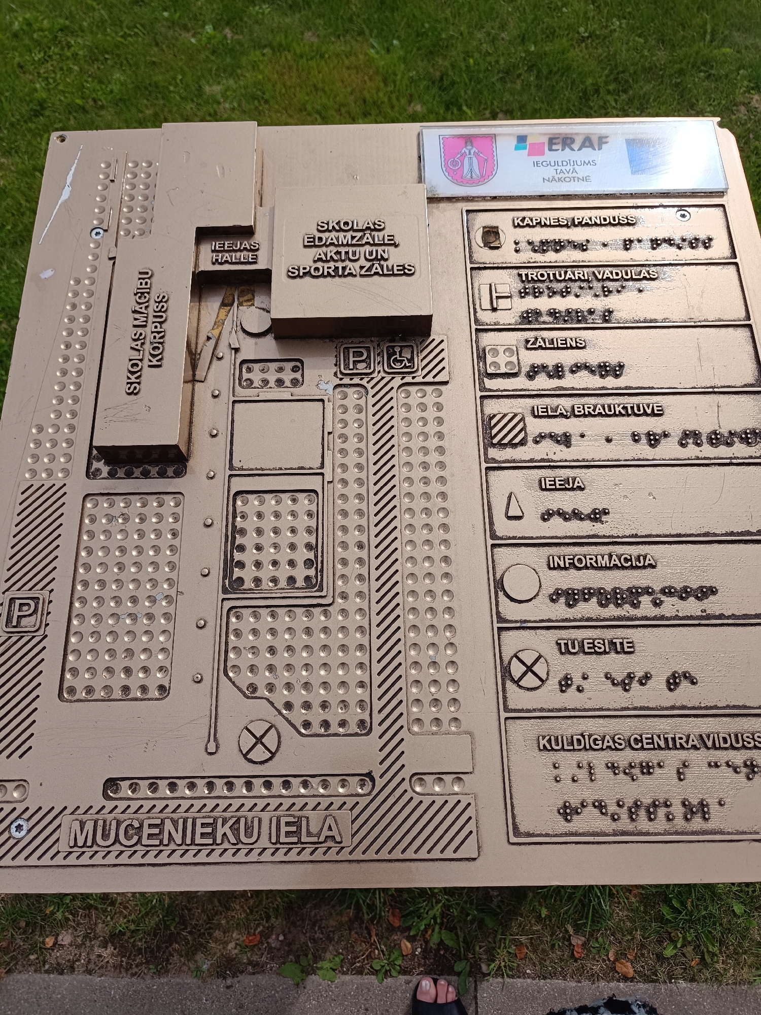 The image shows a tactile map of the Kuldīga Center Secondary School (Kuldīgas Centra vidusskola) in Kuldīga, Latvia. The map is made of a bronze-colored material and features raised text and symbols to aid visually impaired individuals. It depicts the school's main building, labeled "SKOLAS MĀCĪBU KORPUSSS" (School Education Building), and other areas such as the entrance hall ("IEEJAS HALLE"), school dining hall, and sports hall ("SKOLAS EDAMZĀLE, AKTUV UN SPORTA ZĀLES"). The map also includes braille translations of the text and symbols indicating various facilities and directions, such as "KAPNES, PANDUSS" (stairs, elevator), "TROTUĀRI, VADULAS" (sidewalks, ramps), "ZALIENS" (lawn), "IELA, BRAUKTUVĒ" (street, driveway), "IEEJA" (entrance), "INFORMĀCIJA" (information), and "TU ESI TE" (you are here). A section of the map is dedicated to "KULDĪGAS CENTRA VIDUSS" which is the school's name. The map is mounted outdoors, with a grassy area and sidewalk visible in the background. A person's foot wearing a sandal is visible in the bottom left corner of the image.
