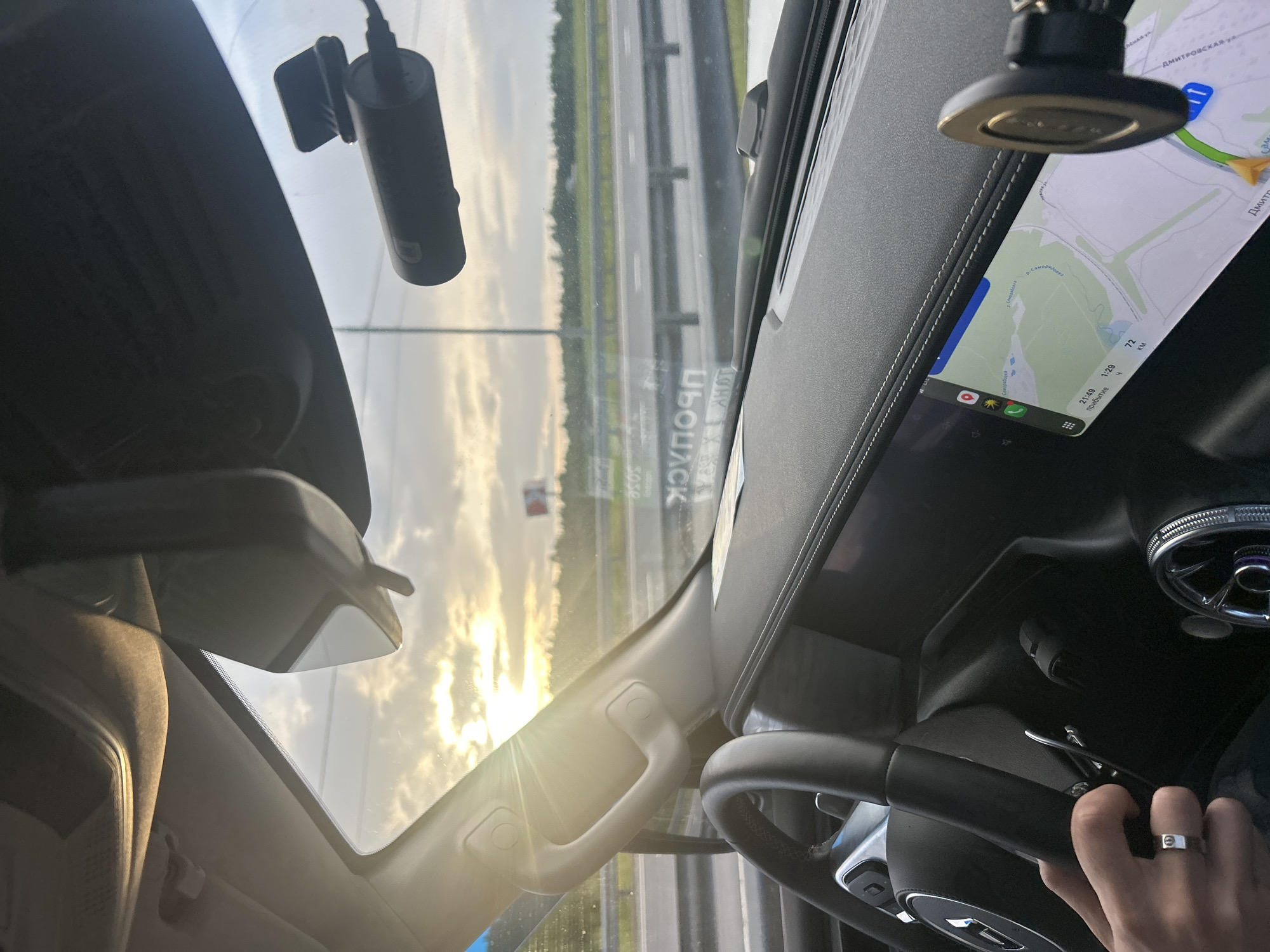 The image shows the interior of a car, likely a modern SUV, as seen from the passenger's perspective. The dashboard features a large central touchscreen displaying a navigation map with the current location identified as Larevo, Russia. The time is 21:49, with an estimated arrival time of 1:29 and a distance of 72 km remaining. A person's hand, wearing a silver ring, is visible on the steering wheel. A dashcam is mounted above the windshield, and a rearview mirror is also present. The windshield offers a view of a highway with trees and cloudy skies with the sun setting. There are also some reflections on the windshield, including a billboard with Russian text.
