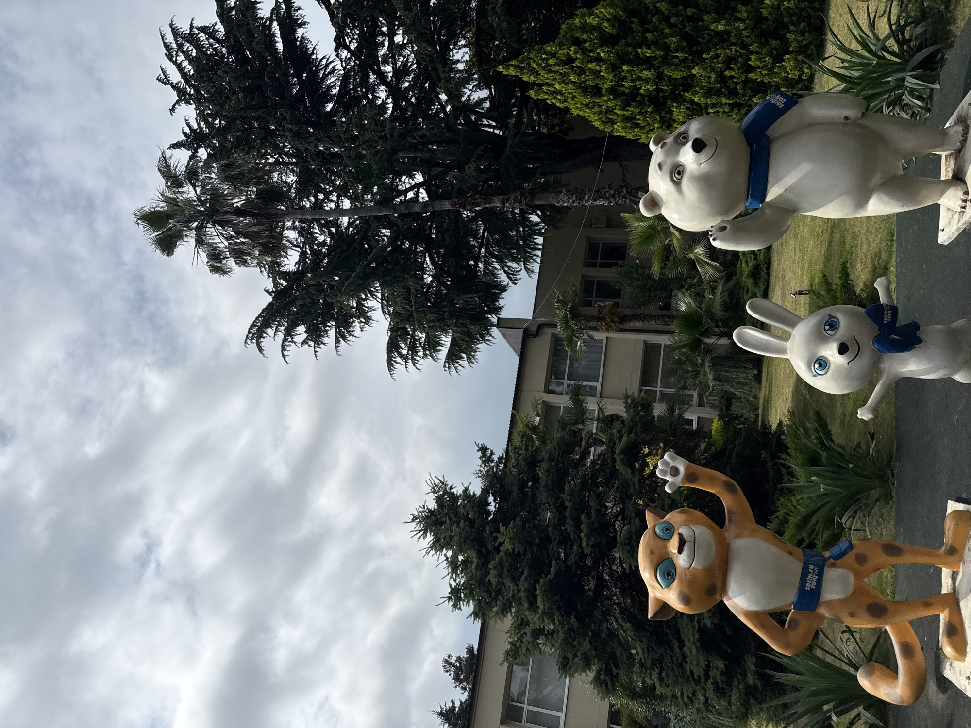 This is a photograph of three Olympic mascot statues, likely from the Sochi 2014 Winter Olympics, displayed outdoors. The statues depict a polar bear, a hare, and a leopard, all in dynamic poses and wearing blue scarves. The polar bear and hare statues are white, while the leopard is orange with brown spots. The scarves on each statue have the text "Sochi 2014" printed on them.
The statues are situated on a gray, rectangular base in front of a building and lush greenery. The building appears to be a light-colored structure with multiple windows. Behind the building, tall trees and palm trees are visible, suggesting a warm climate or a well-maintained park area. The sky is cloudy and overcast, indicating a moderate, possibly cool, weather condition. The overall scene depicts a monument or display commemorating the Sochi Olympics. The statues themselves are cheerful and inviting, conveying a sense of fun and sport. The image is oriented vertically, with the sky and trees occupying the upper portion and the statues and ground in the lower portion.