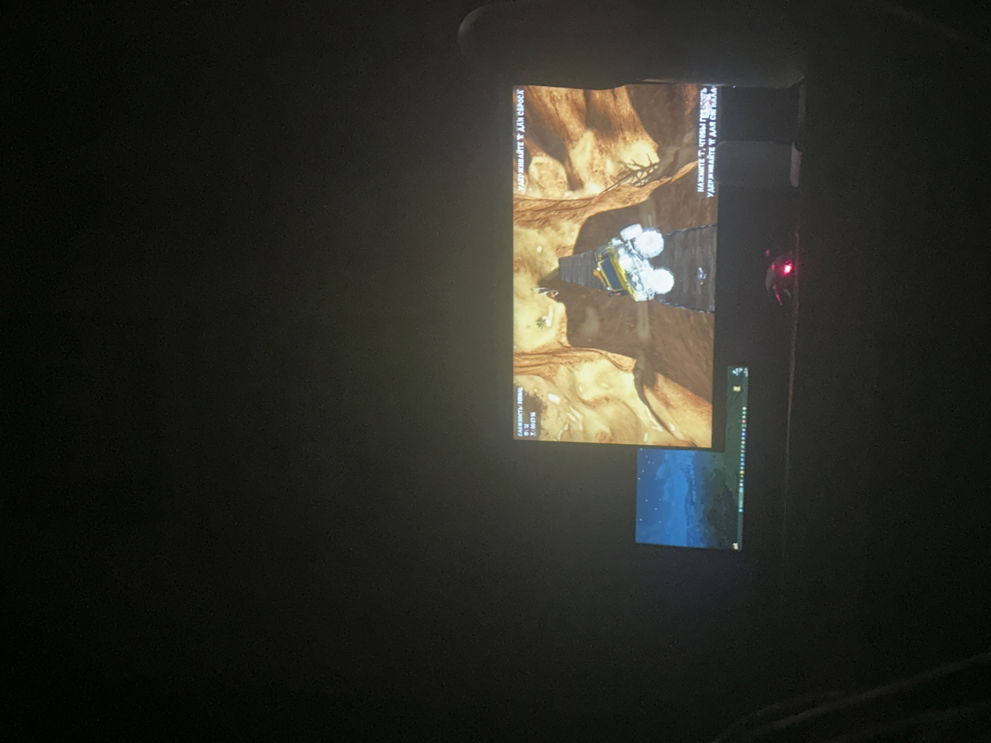 The image depicts a dimly lit room with two computer monitors. The left monitor displays a stylized landscape with a starry night sky. The right monitor shows a video game scene featuring a yellow off-road vehicle navigating a treacherous canyon path. Text overlays on the right monitor, in Russian, read "НАЖИМАЙТЕ Т, ЧТОБЫ ГЕНЕРИРОВАТЬ" (Press T to generate) and "УДЕРЖИВАЙТЕ T ДЛЯ СБРОСА" (Hold T to reset). A red gaming mouse with illuminated buttons is visible on the desk in front of the monitors. The overall setting suggests a gaming session. There are no visible elements that can be used to determine the location of Radošina, Slovakia.