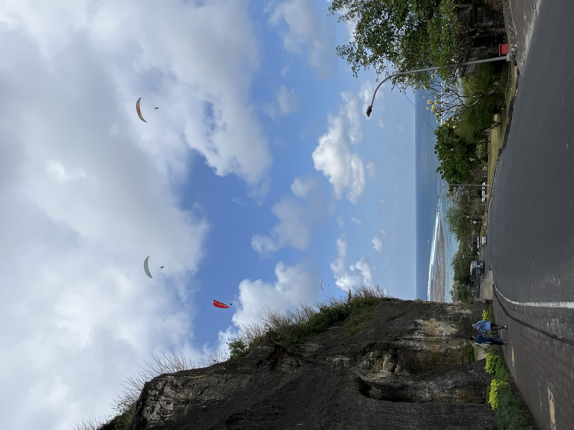 The image depicts a scenic view of the coastline near Kuta Selatan, Indonesia. In the foreground, a paved road curves to the right, bordered by a pedestrian walkway on the left. Two individuals are walking away from the camera on the walkway, on the left side of the road. To the left of the walkway, a sheer cliff face rises, covered in sparse vegetation.
In the sky above, three paragliders are visible. The furthest paraglider, on the right, has a multi-colored canopy and is a significant distance away. A second paraglider, closer to the center, has a light green canopy. The third paraglider, to the left of the center and slightly closer to the cliff, has a red canopy.
Beyond the road and the cliff, the turquoise ocean stretches to the horizon, meeting a bright blue sky dotted with fluffy white clouds. The shoreline features a rocky beach with gentle waves breaking. Further inland, trees and greenery are present, and a few vehicles, including a blue bus, are parked on the side of the road. A lamppost is visible on the right side of the road, near some lush trees and a stone structure. The overall scene is one of natural beauty and recreational activity.