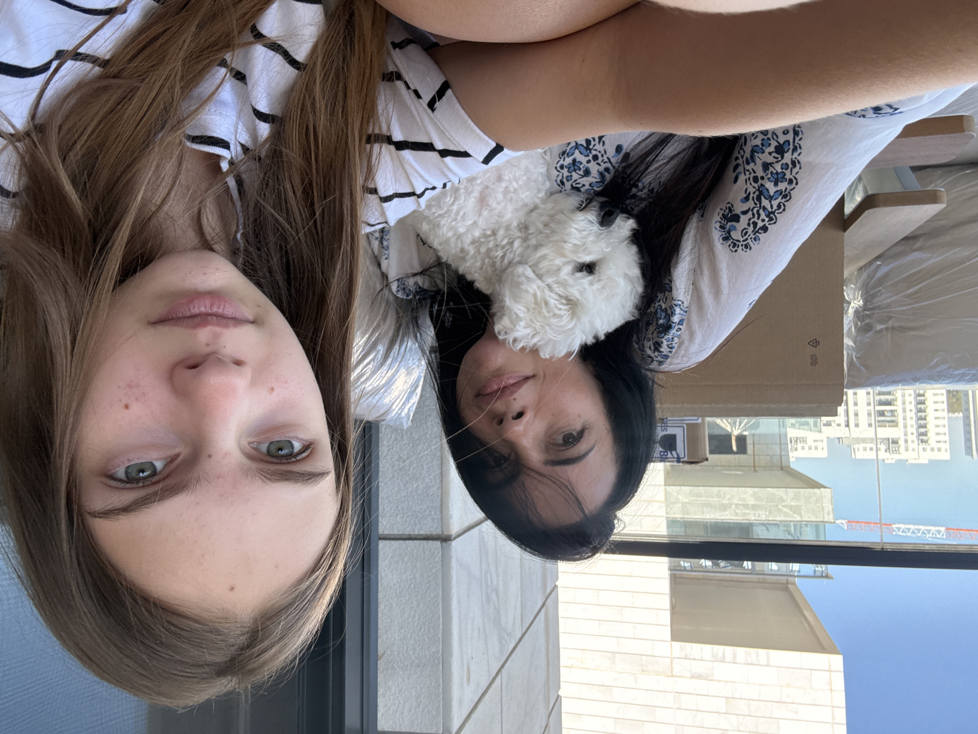 In Or Yehuda, Israel, a woman with dark hair and a white, blue-patterned shirt is holding a small white dog. She is looking towards the camera with a neutral expression. Beside her, a young girl with long brown hair and a white and black striped shirt is also looking at the camera, her gaze directed slightly off-center. The setting appears to be an outdoor balcony, with a modern building and a crane visible in the background, suggesting a city environment undergoing construction. The lighting suggests it is daytime.