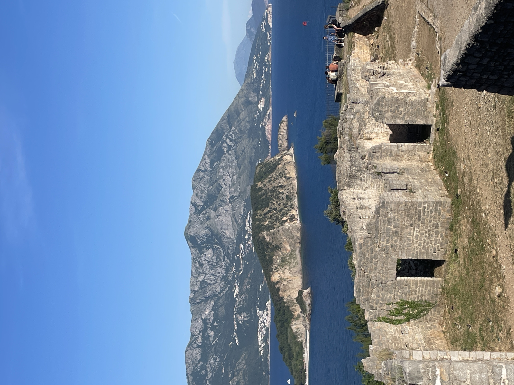 The image depicts a landscape scene from a high vantage point in Budva, Montenegro, overlooking the Adriatic Sea. The sky is clear blue, with mountains in the distance across the sea. The foreground features stone ruins, indicating the remains of a historic structure. Several people are standing near the edge of the ruins, looking out at the view. The sea is dark blue, with a small island or peninsula visible in the middle ground. The ruins are constructed of light-colored stone, with several window openings. Vegetation grows on and around the ruins.