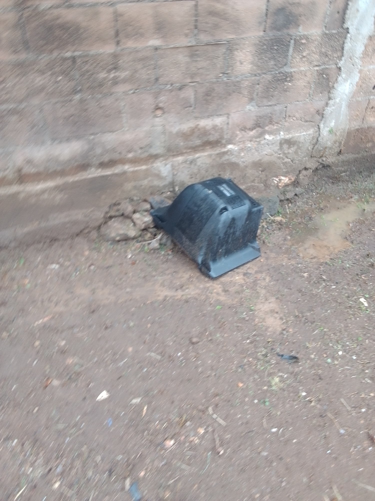 A black, old-fashioned television set lies on its side in the dirt. It appears to be a CRT television, with a bulky casing and a screen that curves inward. The television is resting next to a rough, grey brick wall that shows signs of wear and tear. The ground around the television is made up of dirt and small stones, with some patches of mud and sparse green vegetation. A small puddle of water is visible to the right of the television. The scene suggests the television has been discarded or left outdoors, possibly due to disrepair or being replaced. The lighting in the image is neutral, making it difficult to determine the exact time of day or weather conditions, though the presence of mud and puddles hints at recent rain. The overall setting appears to be an outdoor area, likely a yard or alleyway, with a rustic, unmaintained aesthetic. There is no visible text in the image.