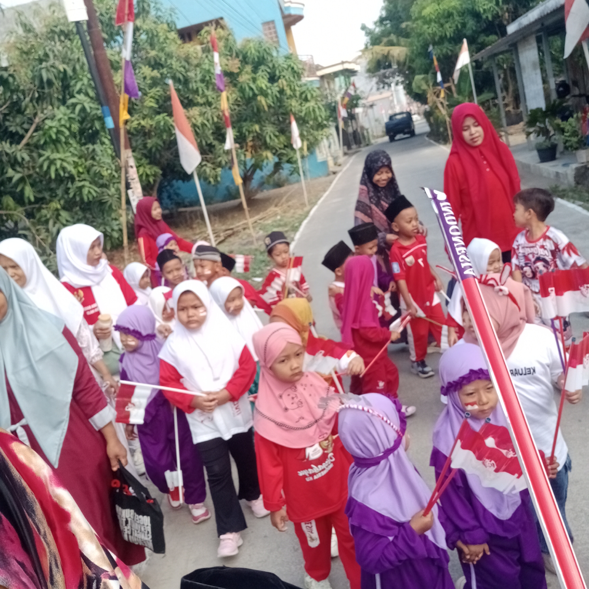 In the city of Tegowanu Kulon, Indonesia, a group of children and adults are gathered outdoors, seemingly for a parade or celebration. Many of the children are dressed in red and white, the national colors of Indonesia, and are holding small Indonesian flags. Some children have face paint. A woman in a bright red hijab stands out among the group. The background features trees and buildings, suggesting a residential or semi-urban environment. The ground appears to be a paved road or pathway. A black car is visible in the distance.