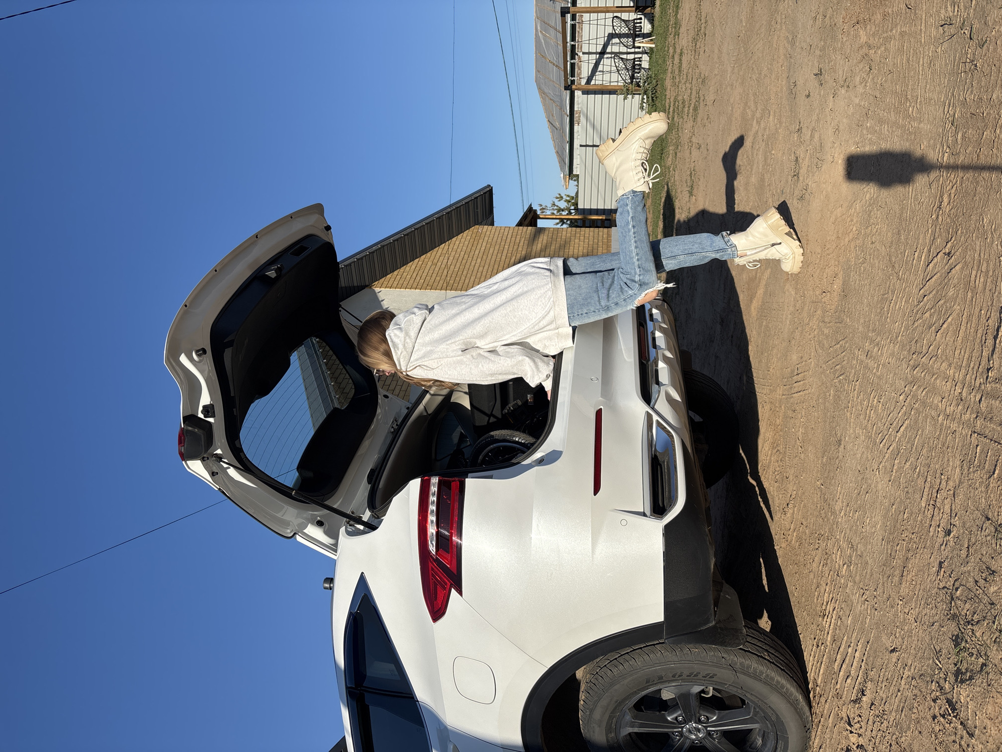 A person is lying in the open hatchback of a white SUV. The person is wearing light blue jeans, a light gray hoodie, and light-colored boots. The vehicle is parked on a dirt or gravel surface. Behind the vehicle is a building with tan siding and a corrugated metal roof. In the background, there is a blue sky. The location is purportedly in the city of Chaikovsky, Russia, though there is no indication of location within the image itself.
