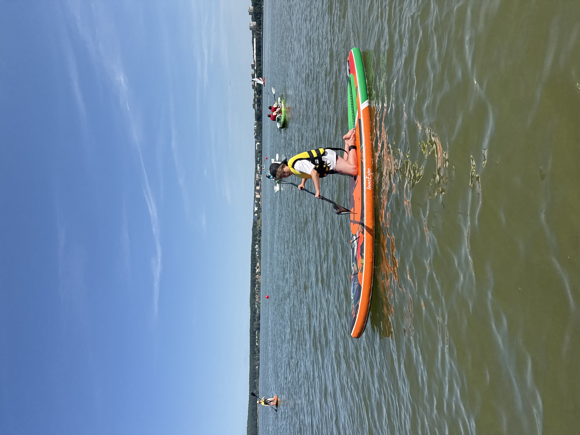 A young person wearing a yellow life vest and a black baseball cap is kneeling on an orange, green, and white paddleboard. They are holding a black paddle in the water and looking towards the camera. The paddleboard has the text "InnoExpo" printed on its side. The person is on a body of water, which appears to be a lake or river, with a wooded shoreline in the background. Several other paddleboards and kayaks are visible in the distance, with people participating in water activities. The sky is clear and blue with a few wispy clouds. The scene is set during the daytime under bright sunlight. This scene is likely located on the Votkinsk Reservoir in Votkinsk, Russia, given the presence of a large body of water, a wooded shoreline, and buildings in the distance that could be part of the city.