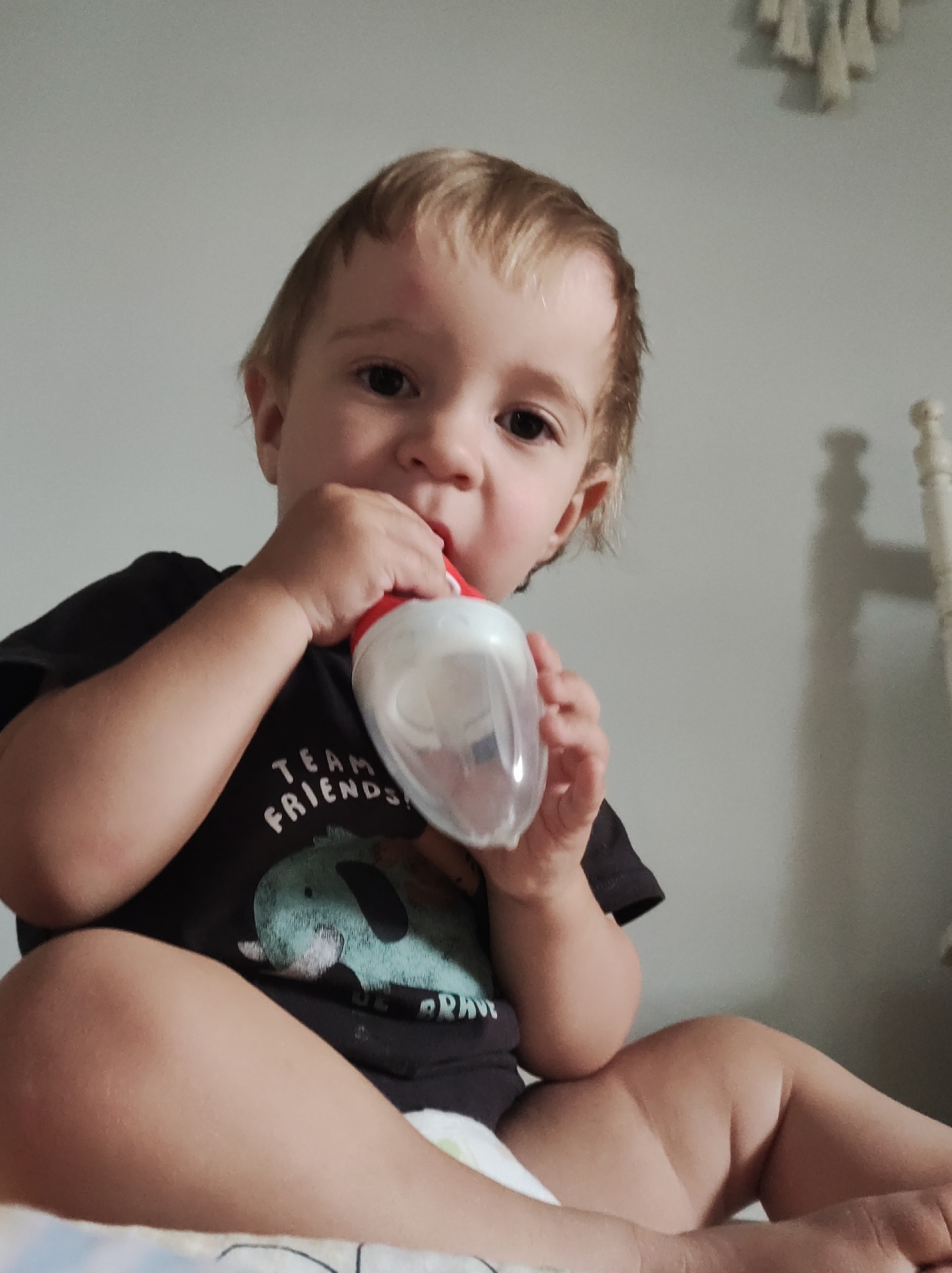 A young child with light brown hair and dark eyes is depicted sitting and drinking from a clear plastic feeding bottle with a red top. The child is looking directly at the camera with a neutral expression. A reddish mark or bump is visible on the child's forehead.
The child is wearing a dark-colored t-shirt with a light blue or green cartoon animal design, possibly an elephant. Visible text on the t-shirt reads "TEAM FRIENDS". The child also appears to be wearing a diaper.
The setting is indoors, with a plain light grey wall serving as the background. On the right side of the frame, part of a white, possibly wooden, bed frame or headboard is visible. Above it, a light-colored, woven or macrame wall hanging can be seen. The lighting suggests daytime, with a soft, even illumination. The child's posture indicates they are sitting on a surface, possibly a bed or couch.