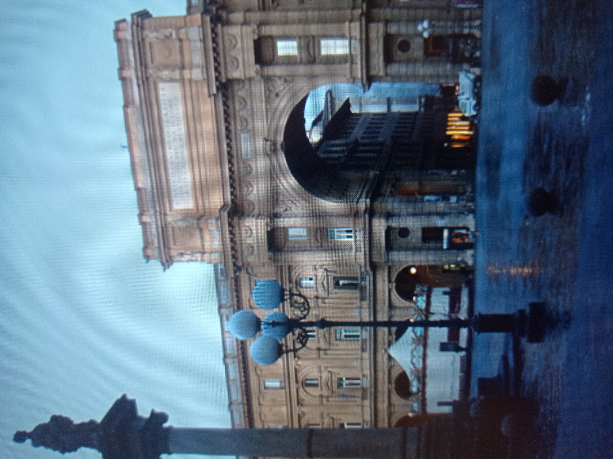 The image depicts a city square with an imposing archway as the central architectural feature. On the left, a tall, dark column topped with a silhouetted statue stands prominently. In the middle ground, a street lamp with three spherical lights is visible. Behind the lamp, a building with an ornate facade, featuring numerous arched windows, is partially obscured. The archway itself is grand, with detailed stonework and columns. Through the arch, another building with a dark facade and lit windows can be seen. In the background, a white tent-like structure is visible. The ground of the square appears wet, reflecting the ambient light. A light-colored vehicle is parked in the distance. The sky is a clear, pale blue, suggesting it is either early morning or late evening. The scene has a muted color palette. The location is identified as the city of Mukachevo, Ukraine.