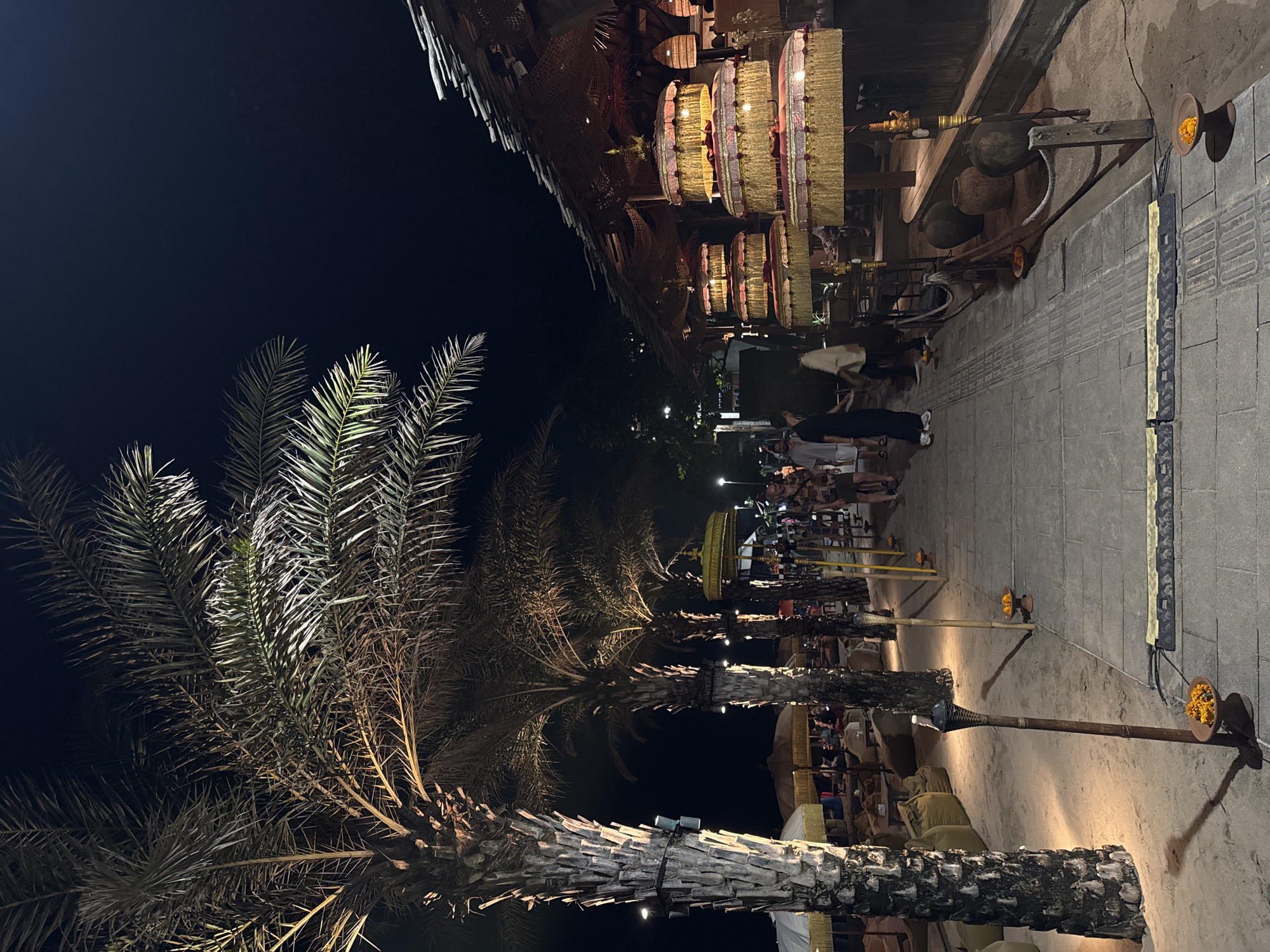 This is a nighttime scene on a beachside walkway in Kuta, Indonesia. The image captures a lively atmosphere with several people walking along the path. On the left, large palm trees with illuminated fronds cast shadows on the sandy ground. A tiki torch stands tall, its light illuminating a small bowl of orange flowers placed at its base. Further down the walkway, several large, tiered umbrellas with gold and red embellishments are displayed, suggesting a commercial establishment or event. The walkway itself is paved with stone tiles, and there are some safety barriers along the edge. In the background, the outline of a thatched-roof building is visible, with lights and lanterns suggesting an open-air restaurant or bar. The overall scene conveys a festive and tropical ambiance characteristic of a popular tourist destination at night.