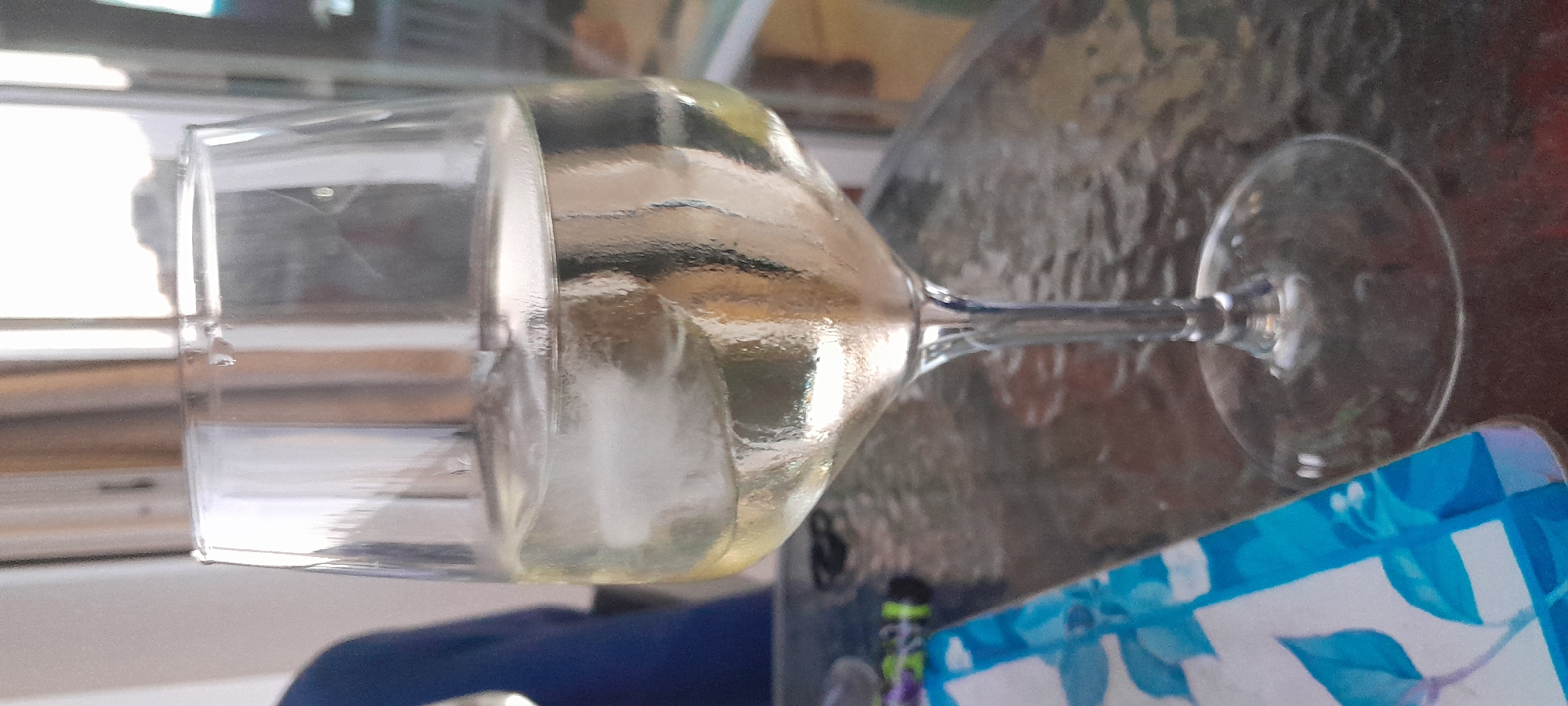 The scene is set in Manilva, Spain. A clear, stemmed wine glass, prominently featured in the foreground, contains a pale yellow liquid with several ice cubes. The glass exhibits minor surface condensation or reflections, indicative of a cold beverage. It rests on a dark, textured glass table surface, possibly a patterned or hammered type, which reflects ambient light. To the immediate left of the glass, a corner of a placemat or napkin with a distinct blue and white floral or leafy pattern is visible, alongside an indistinct, dark object with hints of green and purple. On the far left, a partial view of an individual wearing a blue garment is visible, largely out of focus. The background is brightly lit and heavily blurred, suggesting an outdoor or semi-outdoor setting with large windows or an open vista, reflecting bright sunlight or diffused daylight. The overall composition places emphasis on the beverage and its immediate surroundings.