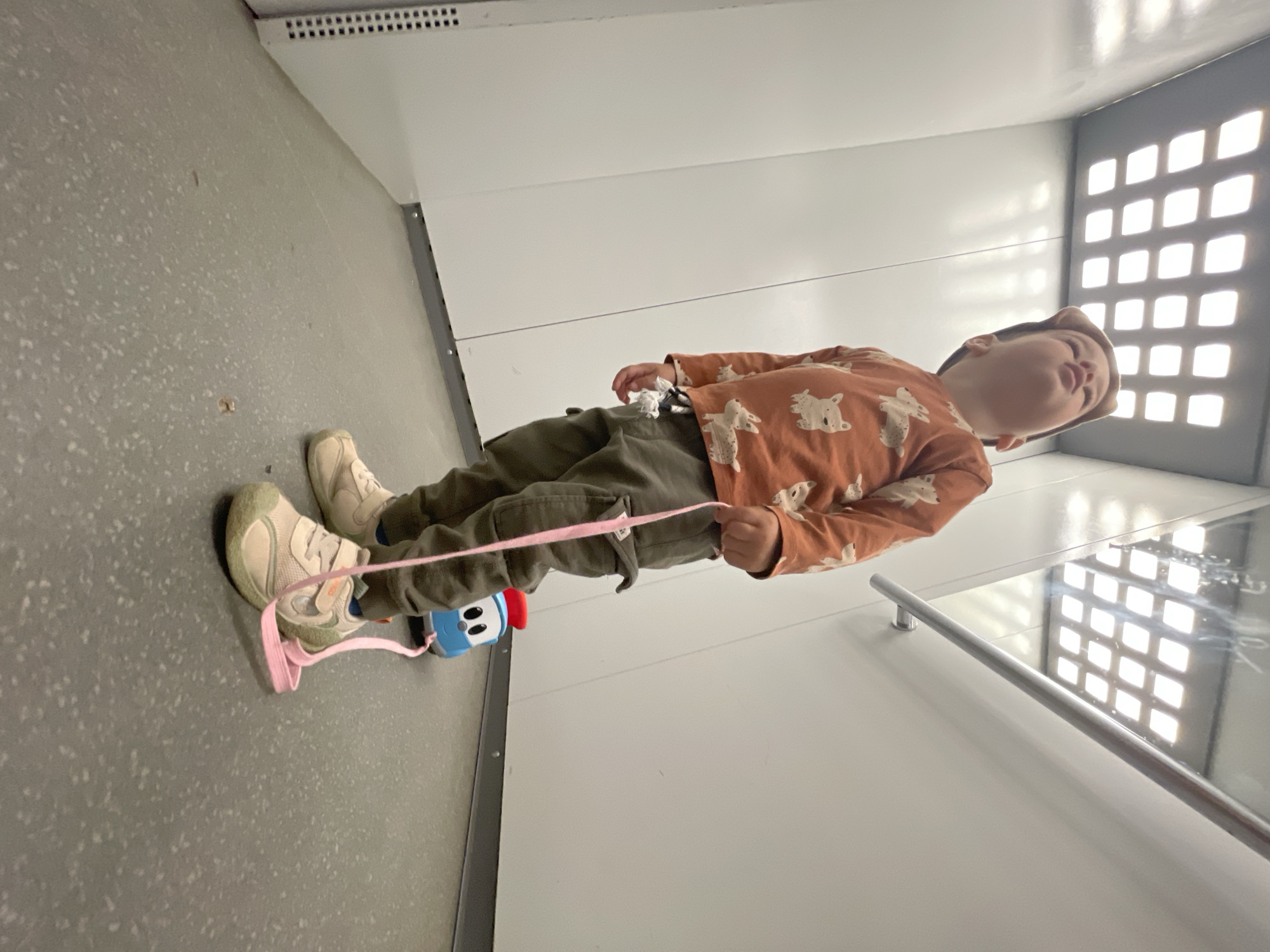 This image captures a young child from a low-angle perspective, standing inside a brightly lit, enclosed interior space. The child is looking directly upwards with a slightly open mouth and a curious expression, towards a grid of approximately 25 recessed, square white lights set into a reflective, light-grey ceiling.
The child is wearing an orange long-sleeved shirt adorned with multiple white dog or animal patterns, olive green cargo-style pants, and light-colored (beige/tan) athletic shoes with velcro straps. A brown cap, possibly with decorative ears, is on the child's head. The child's right hand is holding a pink fabric strap or leash which extends downwards, seemingly connected to a small blue toy car with a white face, black eyes, and a red cap or light on its top, positioned behind the child's left leg.
The environment features smooth, unadorned white wall panels. On the right side, a metallic horizontal handrail is affixed to the wall, reflecting the overhead lights. On the lower left, a ventilation panel with a pattern of small square perforations is visible. The floor is a textured, speckled grey material, showing a few small brown specks of debris.
Based on the architectural elements—the grid ceiling lights, reflective surfaces, smooth walls, handrail, and confined nature—the scene is most consistent with the interior of an elevator, a modern public transport vehicle (such as a tram or train car), or a minimalist building lobby in Izhevsk, Russia. The specific address or building within Izhevsk cannot be determined from the visual information provided.