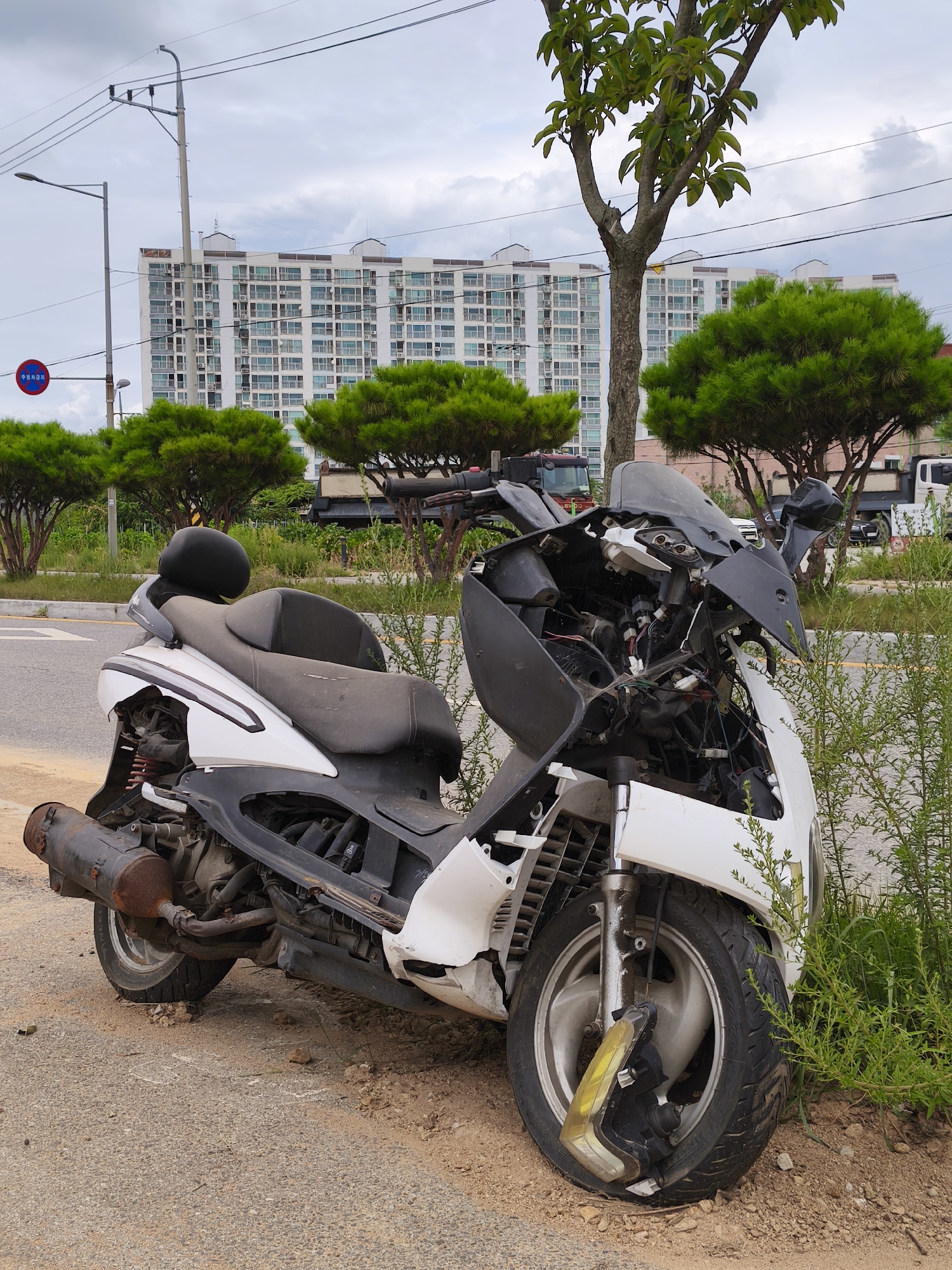 A damaged white scooter is parked on the side of a street in Asan-si, South Korea. The scooter is positioned at an angle, with its front wheel turned slightly to the left and resting on the unpaved shoulder. The front fairing is extensively damaged, with exposed wiring and broken plastic components. The headlight is detached and hanging precariously. The exhaust pipe on the left side is rusty and discolored. The body of the scooter, particularly the white panels, appears dirty and worn. The seat is a dark color, possibly black, and also shows signs of wear and dust. The surrounding environment includes a paved road to the left, a gravel and dirt shoulder, and green vegetation, including some bushes and trees. In the background, there is a tall, modern apartment building with many windows. Power lines and utility poles are visible against a cloudy sky. The overall scene suggests a neglected or abandoned vehicle.
