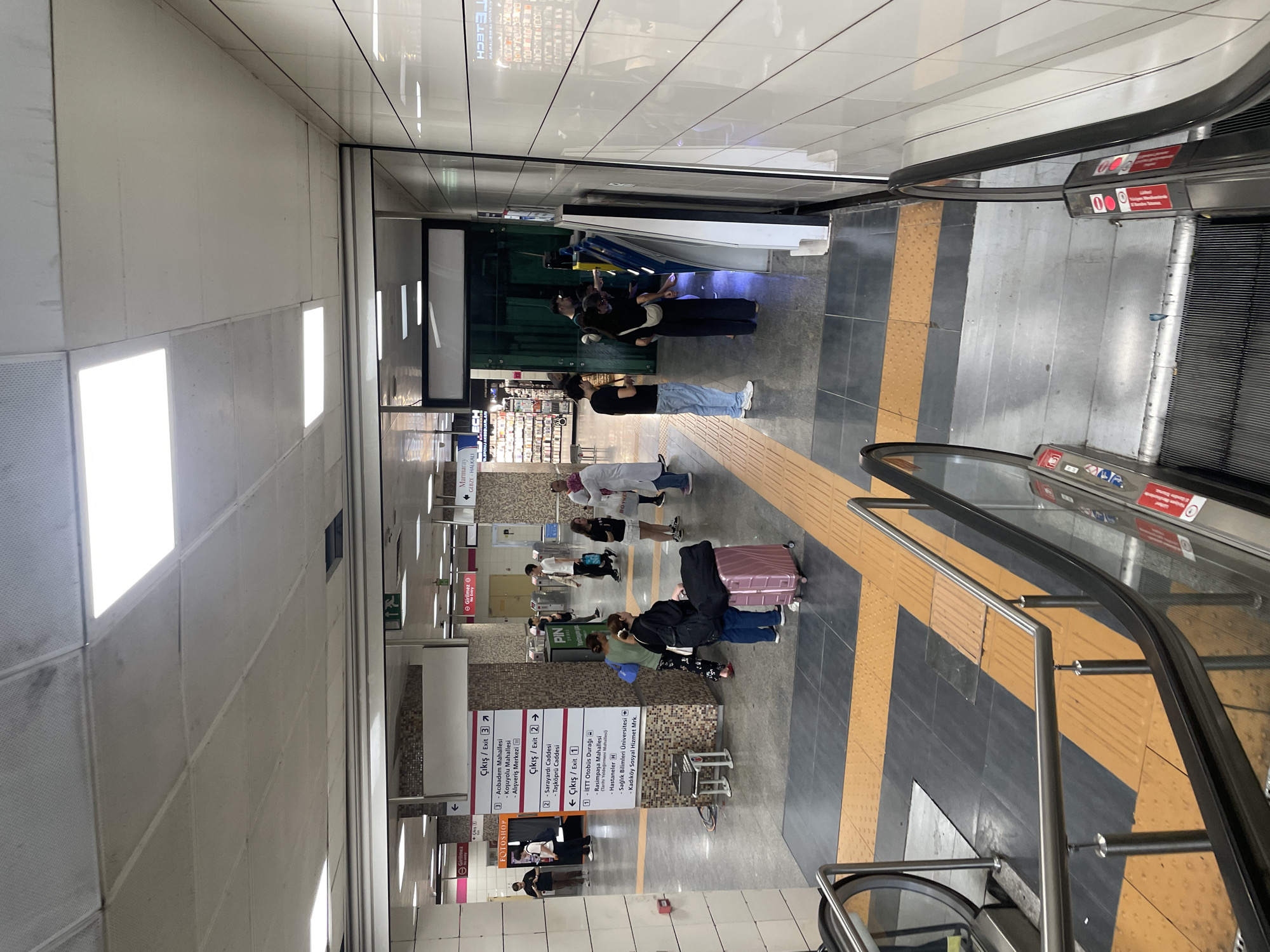 The image captures a scene inside the Kadıköy Metro Station in Istanbul, Turkey, an underground transit hub. In the foreground, a set of escalators descends, their metallic railings and steps visible. To the right, a white tiled wall with a reflective surface lines the station. In the middle ground, a group of people are interacting with ticket machines. One person, wearing a black top and pants, stands closest to a yellow and blue machine, possibly making a purchase. Another person, in a black t-shirt and blue jeans, stands to their left. Further back, several individuals are walking through the station. To the left, directional signage is posted, indicating exits and nearby locations in Turkish, such as "Çıkış / Exit" and station names. One sign lists various destinations including "Acıbadem Mahallesi," "Koşuyolu Mahallesi," and "Hastaneler" (Hospitals). In the background, shops are visible, including a "FOTO SHOP." The overall environment is typical of a busy urban subway station, with fluorescent lighting illuminating the tiled floors and walls.