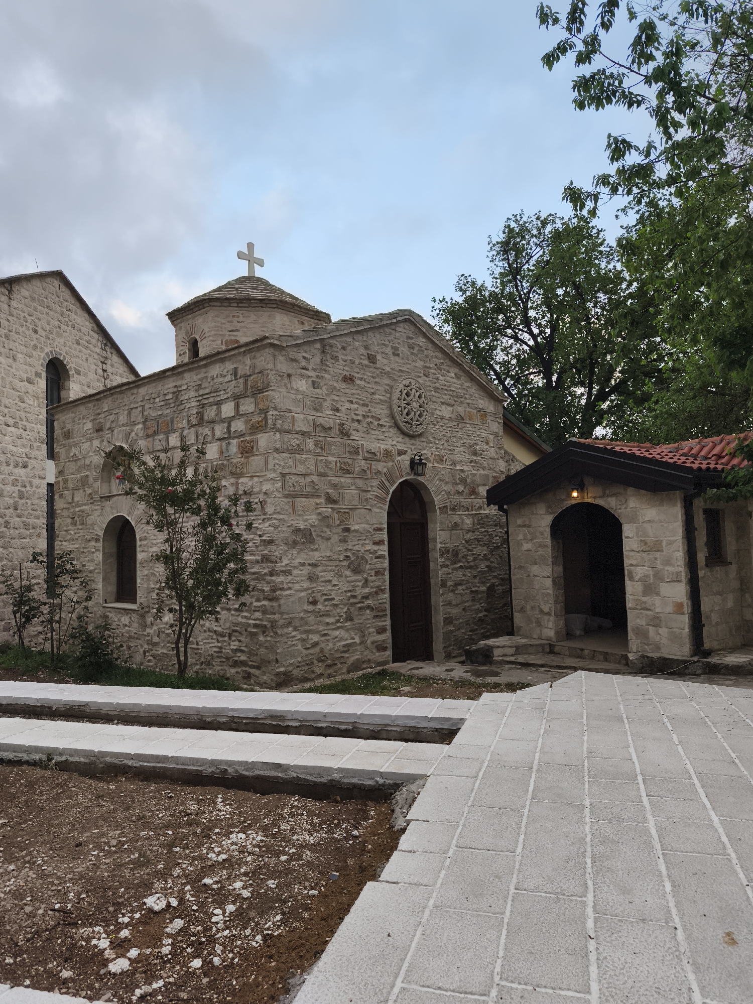 This image captures a historical stone church complex, likely a monastery, set against an overcast sky in Budva, Montenegro. No people are visible in the scene.
The central subject is a robust stone building constructed from irregular, light-colored blocks. It features a prominent dome topped with a white cross. Below the dome, there are small arched openings. The main facade of this building includes an arched wooden door and a circular stone carving above it, possibly an ornamental rose window. To the left, an adjoining, taller stone structure with similar architectural elements and arched windows is visible. To the right, a smaller, separate stone annex or porch with a red-tiled roof and an arched entrance stands beside the main church. This smaller structure has a traditional lantern light fixture above its entrance.
The foreground consists of newly laid light-colored stone slab pathways, appearing as if recent construction or renovation has taken place. Between these pathways and leading up to the buildings, the ground is bare, disturbed earth with scattered white pebbles, suggesting ongoing landscaping. Several young trees and bushes are planted near the buildings. In the background and to the far right, lush green foliage from mature trees frames the architectural complex, indicating a natural, perhaps secluded, setting.
The time of day appears to be daytime, possibly late afternoon or an overcast day, given the soft, diffused lighting and the partially cloudy sky with hints of blue. The overall atmosphere is one of tranquility and historical reverence. No visible text is present in the image.