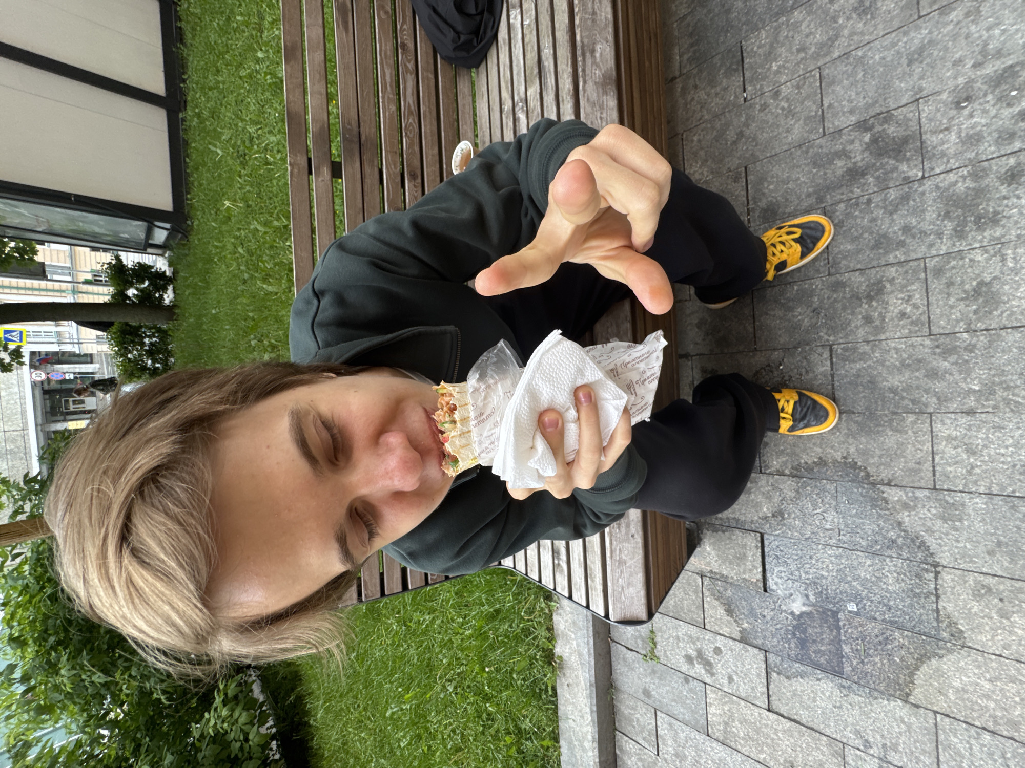 A person is sitting on a wooden bench in what appears to be a city park or public space. They are eating a shawarma or similar wrap, holding it with one hand and gesturing with the other. The person is wearing a dark green hoodie and black pants with yellow and black sneakers. The ground is paved with grey rectangular stones. The location is identified as Moscow, Russia. In the background, there is a green lawn and some buildings visible across the street.