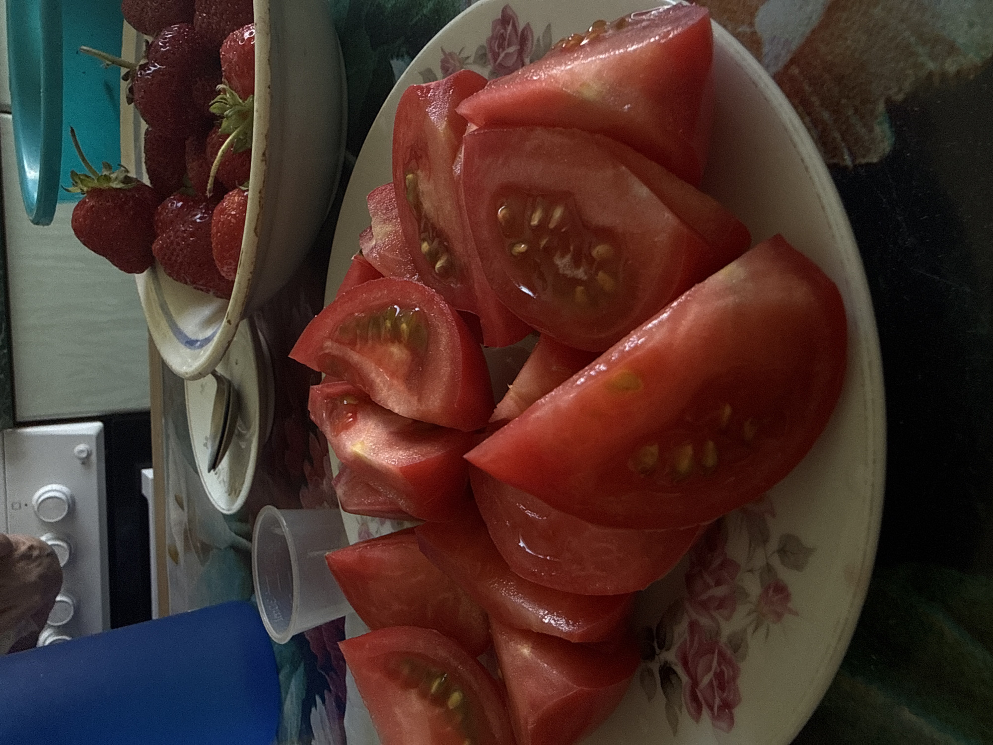 This media file captures a close-up, angled view of chopped tomatoes arranged on a floral-patterned plate. The tomatoes are bright red and appear freshly cut, with visible seeds and juice. To the left of the plate, a portion of a bowl filled with strawberries is visible. The strawberries are a deep red and also appear fresh.
The background is somewhat cluttered, showing elements that suggest a kitchen setting. A white appliance with circular knobs is partially visible on the left, along with a blue cylindrical object and a clear plastic container. The overall lighting suggests it could be daytime, but the specific time of day is not clearly discernible. There are no clear indicators of weather or specific emotions.
The image is a still shot, likely taken in a domestic environment. The presence of fresh produce like tomatoes and strawberries indicates the preparation of food or a meal. No discernible text is visible in the image. The location context is Dnipro, Ukraine, but this cannot be visually confirmed from the image itself.