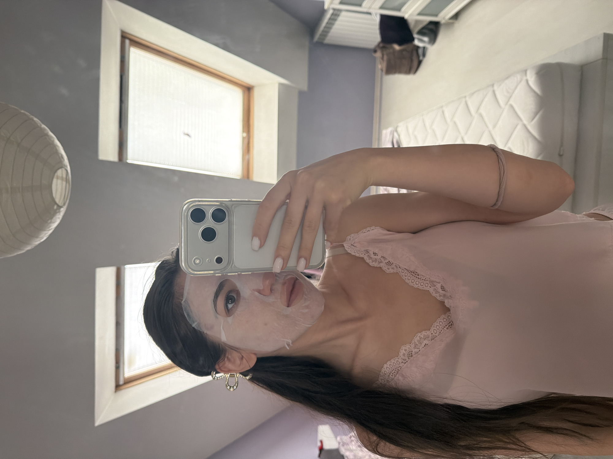 This media file is a portrait-oriented, vertical, slightly angled selfie taken in what appears to be a bedroom. The subject is a young woman with long, dark hair and a sheet face mask covering her face. She is wearing a light pink lace-trimmed camisole. She is holding a smartphone in her right hand, which is angled towards the mirror to capture the reflection.
The background reveals elements of a room with sloping ceilings, characteristic of an attic or top floor. Two windows with white blinds are visible on the left side. On the right side of the reflection, a white mattress on a bed frame and some shelves with clothing or bags can be seen. A large, white, circular paper lantern hangs on the left side of the actual room. The lighting suggests it might be daytime, with natural light coming through the windows.
The woman's expression is neutral, and she appears to be engaged in a routine skincare activity. The overall mood is casual and personal, suggesting a moment of self-care at home. There is no visible text in the image. The location context provided is Sofia, Bulgaria.