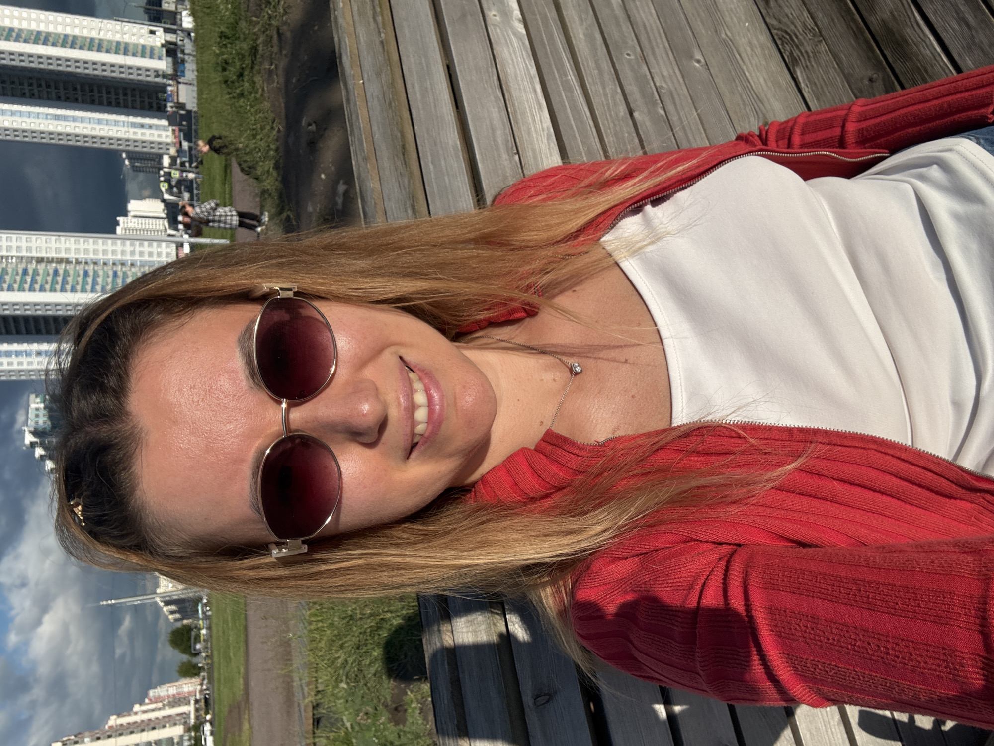 A medium shot captures a Caucasian woman in her late 20s or early 30s sitting on a wooden bench in an outdoor urban setting. She has long blonde hair with some highlights, pulled back into a ponytail with a clip. She is wearing large, oval-shaped sunglasses with a rose-gold colored frame and lenses that appear to be tinted red. Her attire consists of a white tank top and a bright red, zip-up cardigan. A delicate silver necklace with a small, clear pendant is visible around her neck. The woman is smiling and looking directly at the camera.
The background shows a paved area leading to a grassy expanse, with several tall, modern buildings in the distance. The sky is partly cloudy, with dramatic dark clouds present. To the right of the woman, a shadow of a person or object is cast on the wooden bench. In the far background, two people, one wearing a plaid jacket and the other in darker clothing, can be seen walking in the distance. The overall lighting suggests it is daytime, with the sun casting some shadows. The environment appears to be a park or recreational area within a city.