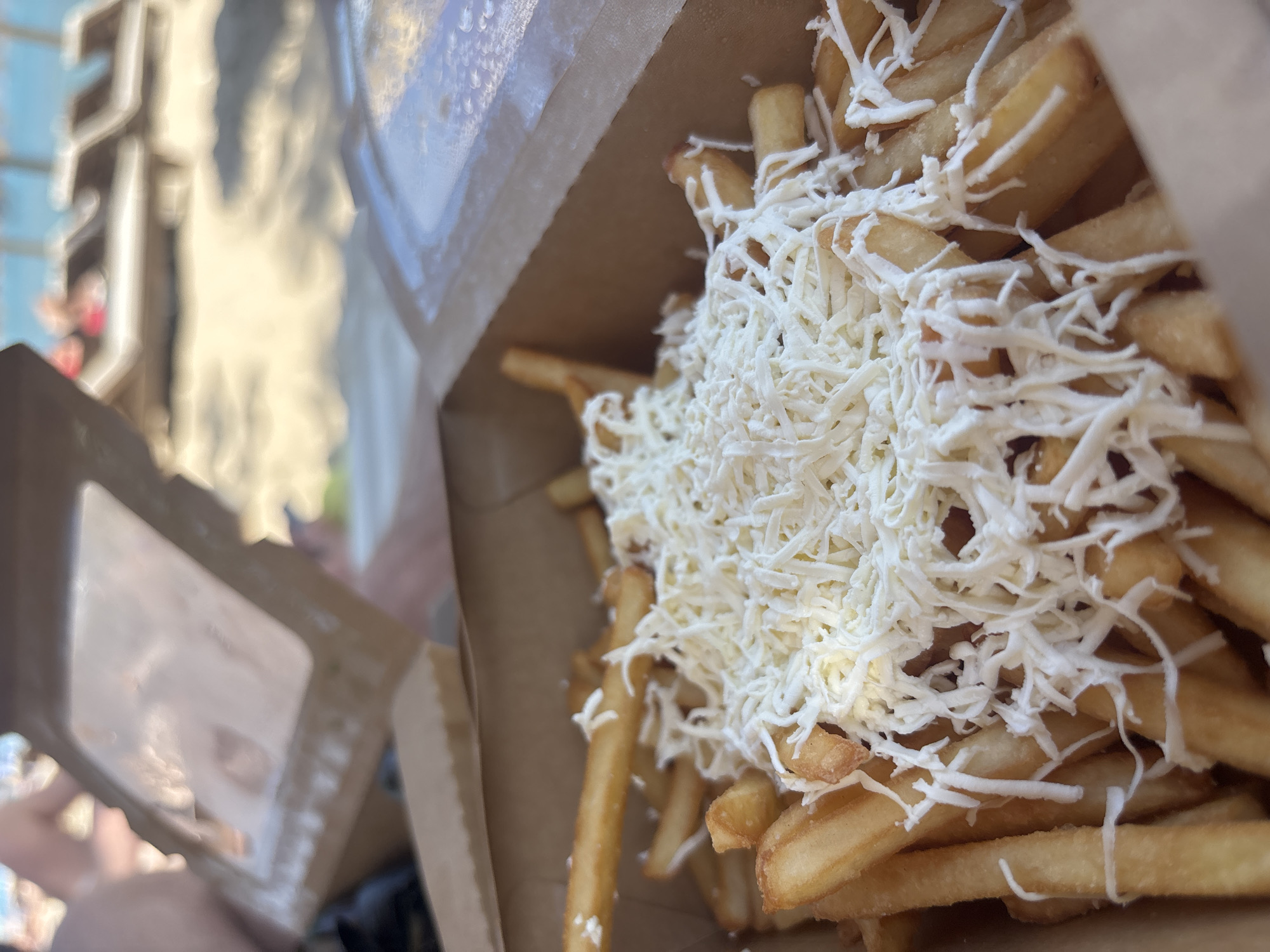 A close-up image presents golden-brown french fries extensively covered with shredded white cheese, served in an open, light brown cardboard take-out container. The cheese remains largely unmelted. In the blurred midground, portions of additional similar take-out containers and indistinct human forms are visible. The background is deeply out of focus, revealing a sandy beach featuring multiple lounge chairs, some occupied by blurred individuals. Beyond the sandy area, a body of water, consistent with the Black Sea, is discernible under bright daylight conditions. The scene depicts a beach environment in Kranevo, Bulgaria.