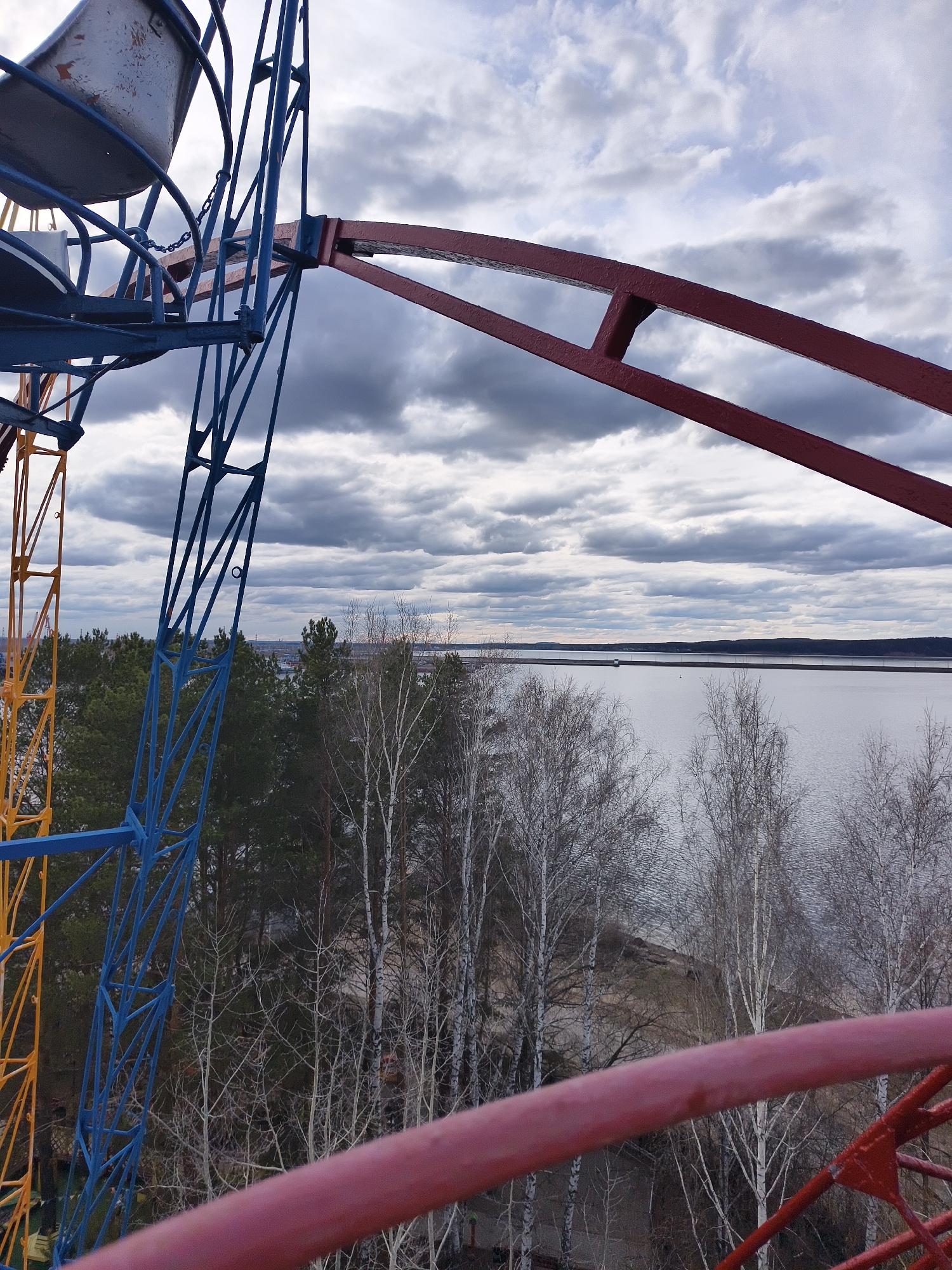 This image presents a high-angle view from an elevated structure, likely a Ferris wheel, overlooking a vast natural landscape under an overcast sky. In the immediate foreground and extending into the mid-ground, sections of the large metal structure are prominent. These include blue, yellow, and red painted girders and beams. A weathered, off-white bucket seat, characteristic of an amusement park ride, is visible in the upper left corner, attached to the blue framework by chains.
The natural scenery unfolds beyond the metallic framework. A dense band of trees occupies the mid-ground, featuring dark green conifers mixed with numerous deciduous trees, primarily tall birches, which are bare of leaves, suggesting late autumn, winter, or early spring. Beyond the trees, a wide expanse of calm water, possibly a lake or reservoir, dominates the horizon. A narrow strip of land or a distant dam/causeway stretches across the water. The sky above is heavily clouded with varying shades of grey, indicating an overcast or cloudy day. No people are visible, and there is no discernible text in the image. The overall impression is one of an expansive, somewhat stark landscape viewed from an industrial-style recreational structure.