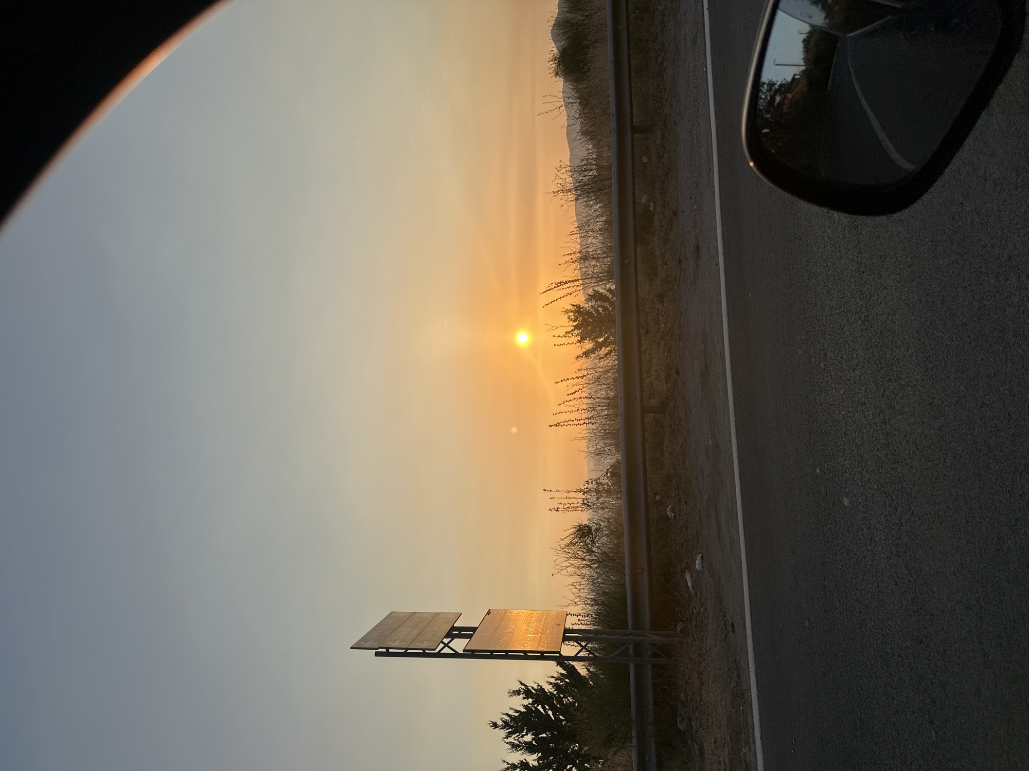 The image is a scenic view of a road at sunset, possibly near Sveti Stefan, Montenegro. The sun is low in the sky, casting a warm orange glow across the horizon. In the foreground, a dark asphalt road is visible, with a white line marking the edge. A car's side mirror is partially visible on the right side of the frame, reflecting the road and some trees.
To the left of the road, there is a metal guardrail separating the road from a grassy embankment. On the embankment, a tall metal pole with two rectangular signs is planted. The signs appear to be directional, with some text and arrows on them, illuminated by the setting sun. Behind the guardrail, dry grasses and some sparse trees are seen. Further in the background, rolling hills are silhouetted against the colorful sky. The overall impression is one of a quiet, rural road bathed in the soft light of dusk. There are no people or animals visible in the image.