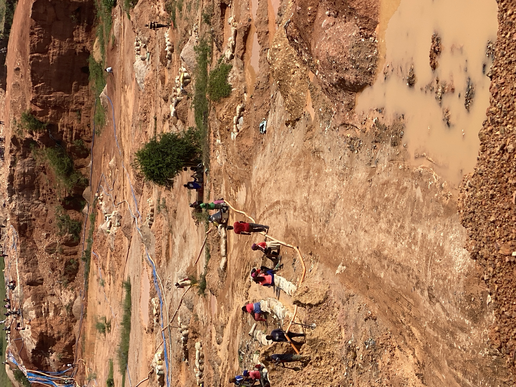 The image depicts an active, open-pit mining operation in the city of Jos, Nigeria. The landscape is characterized by rough, reddish-brown earth, with visible excavations and piles of excavated material. Water, likely from recent rainfall or the mining process itself, has formed muddy puddles and channels throughout the site.
Several individuals are engaged in various activities related to the mining. Some are digging with shovels, while others appear to be transporting materials, possibly using wheelbarrows or carrying sacks. There are also groups of people gathered, potentially taking a break or discussing work. Orange hoses, likely used for water transport or extraction, are visible across the site. The presence of sandbags suggests efforts to manage water or stabilize the terrain.
The overall impression is one of manual labor in a challenging and dynamic environment. The terrain is uneven, with steep inclines and depressions, indicative of ongoing excavation. The color palette is dominated by earth tones, with the vibrant colors of some individuals' clothing providing points of contrast. The scene suggests a busy, yet potentially hazardous, working environment.