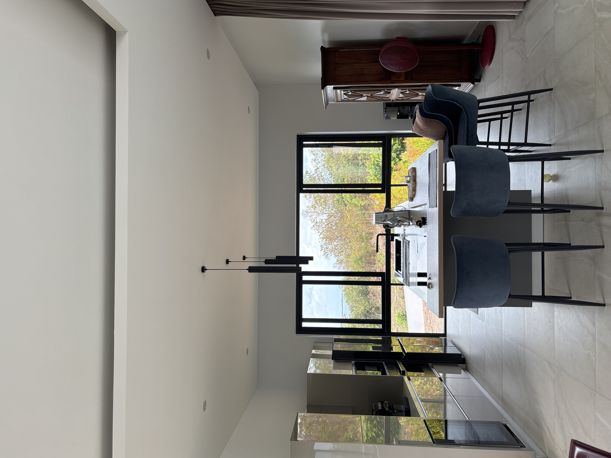 The image shows the interior of a modern kitchen in a location presumed to be in Ban Bang Khao, Thailand, based on inference. The dominant features include: a kitchen island with a light-colored countertop and gray cabinetry, three bar stools with dark blue upholstery and black legs positioned at the island; a stainless steel appliance unit with a microwave and oven; a set of pendant lights suspended above the island; and a large window offering an exterior view of trees. The room is illuminated by natural light. At the far end of the room is a wooden cabinet, upon which sits a red bowl. The floor appears to be tiled. A beige curtain is also seen. The space is generally uncluttered.