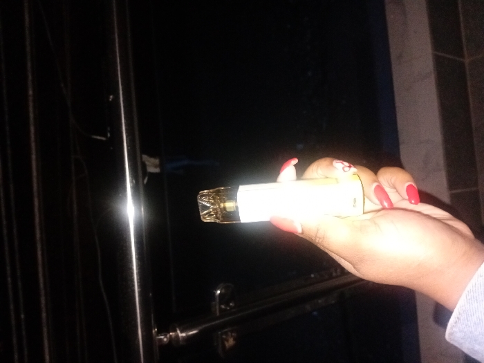 A hand holds a vape pen against a dark background. The hand has a light complexion and sports red manicured nails. The vape pen is illuminated, revealing a clear mouthpiece and a lighter-colored body. A metal gate with vertical bars is faintly visible to the left. The overall scene is dimly lit. The photo appears to be taken at night, focusing on the hand and the device.