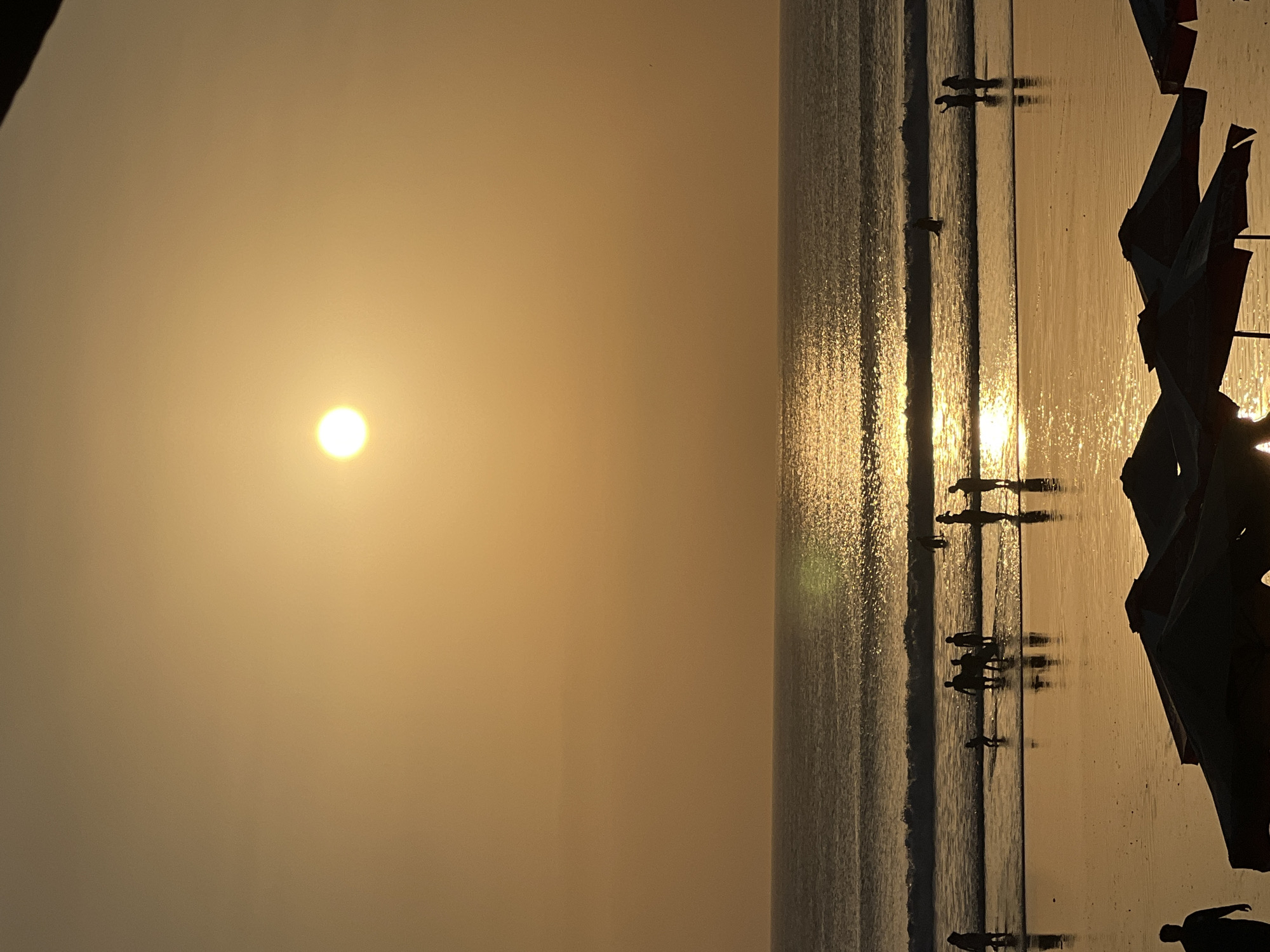 The image captures a serene sunset scene at Kuta Beach, Indonesia. The sun, a bright, diffused orb, hangs low in the sky, casting a warm, golden glow over the entire panorama. Its reflection shimmers intensely on the surface of the ocean, creating a luminous pathway that stretches towards the shore. Gentle waves roll onto the beach, their foamy crests catching the light.
Silhouettes of people are visible wading in the shallows, their forms indistinct against the bright backdrop. Some are standing, while others appear to be walking or playing in the water. A few individuals are gathered in small groups, suggesting casual social interaction. Further up the beach, the dark shapes of beach umbrellas are scattered, some open and others closed, hinting at a typical beach day winding down. The wet sand reflects the golden light of the sunset, mirroring the figures and the umbrellas. The overall atmosphere is one of peaceful twilight, with the natural beauty of the sunset dominating the scene.
